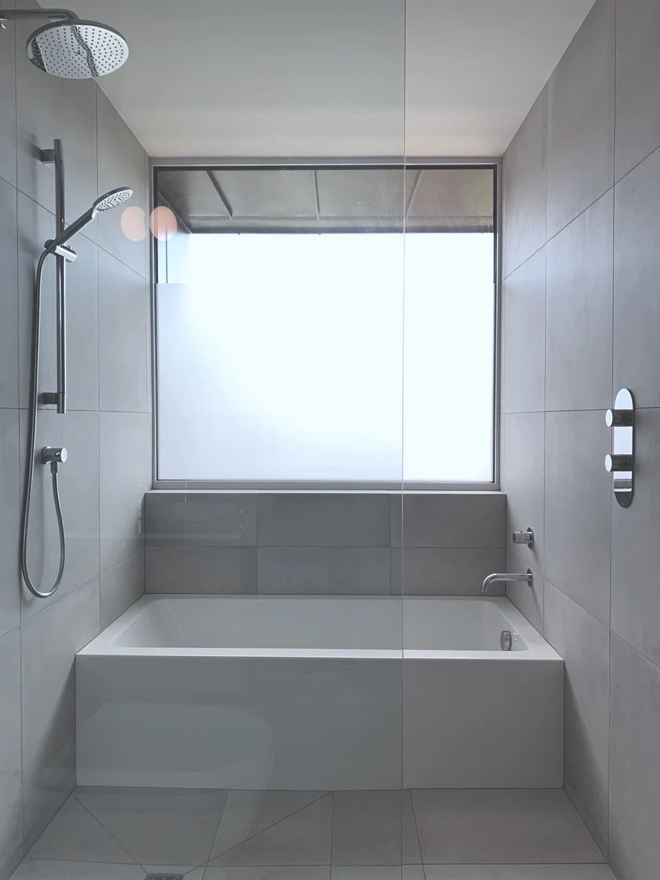 Modern bathroom with a built-in bathtub, gray tiled walls, a large frosted window, and a walk-in shower with a rainfall head.