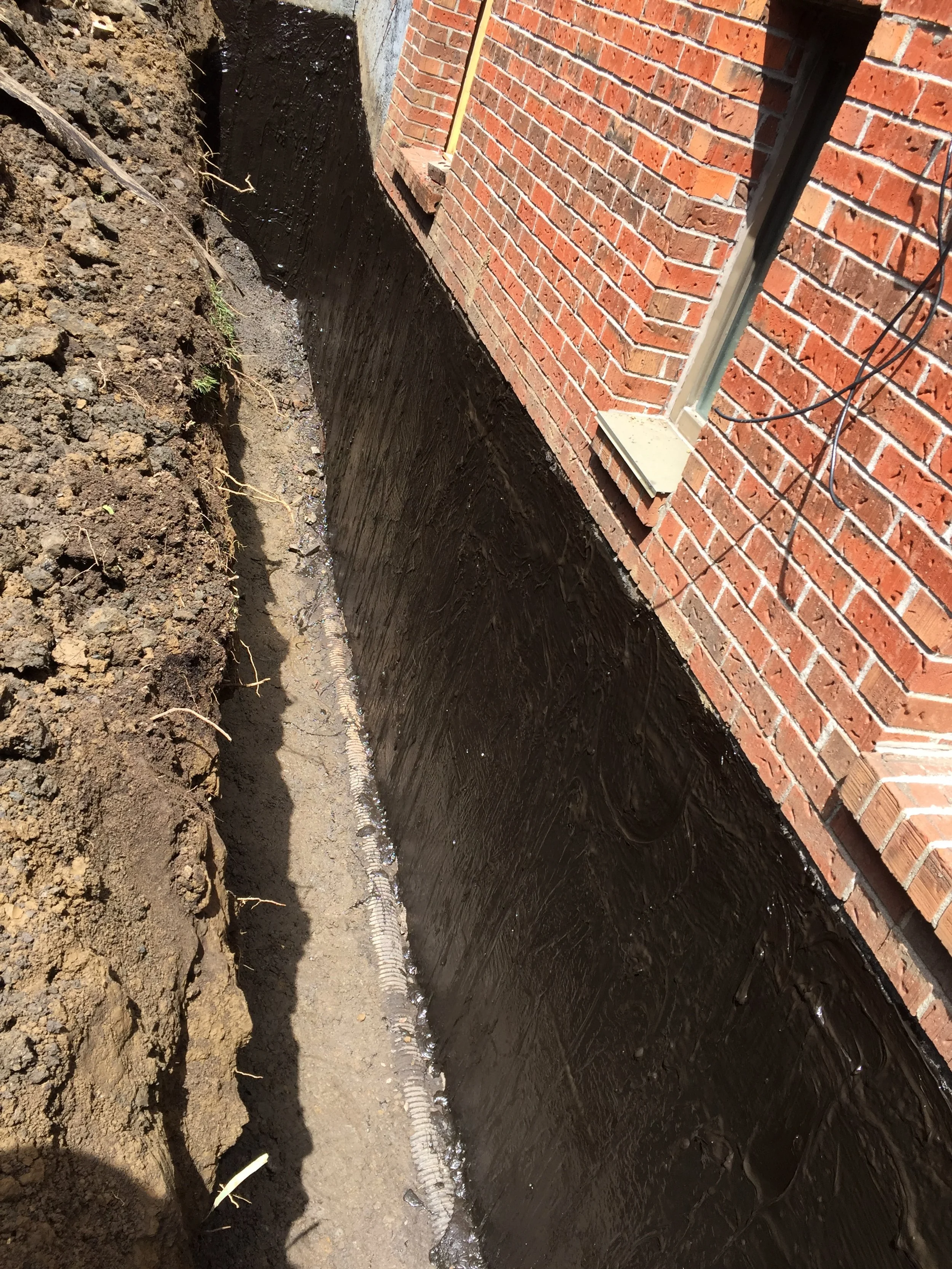 Excavation site next to a brick house foundation with black waterproofing membrane installed on the exterior wall, showing a deep trench with soil on one side.