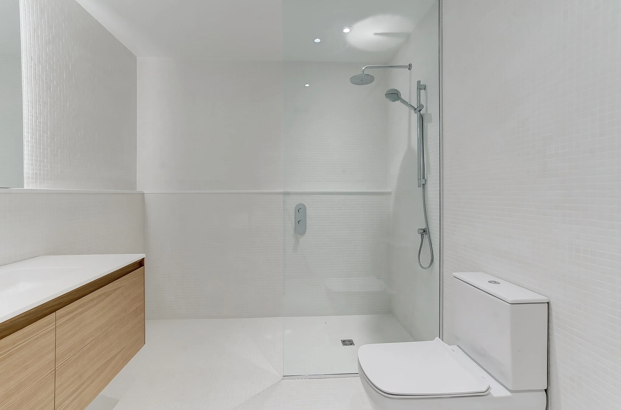 Minimalist bathroom with white tiles, a wooden vanity with a white countertop, a walk-in shower with glass partition.