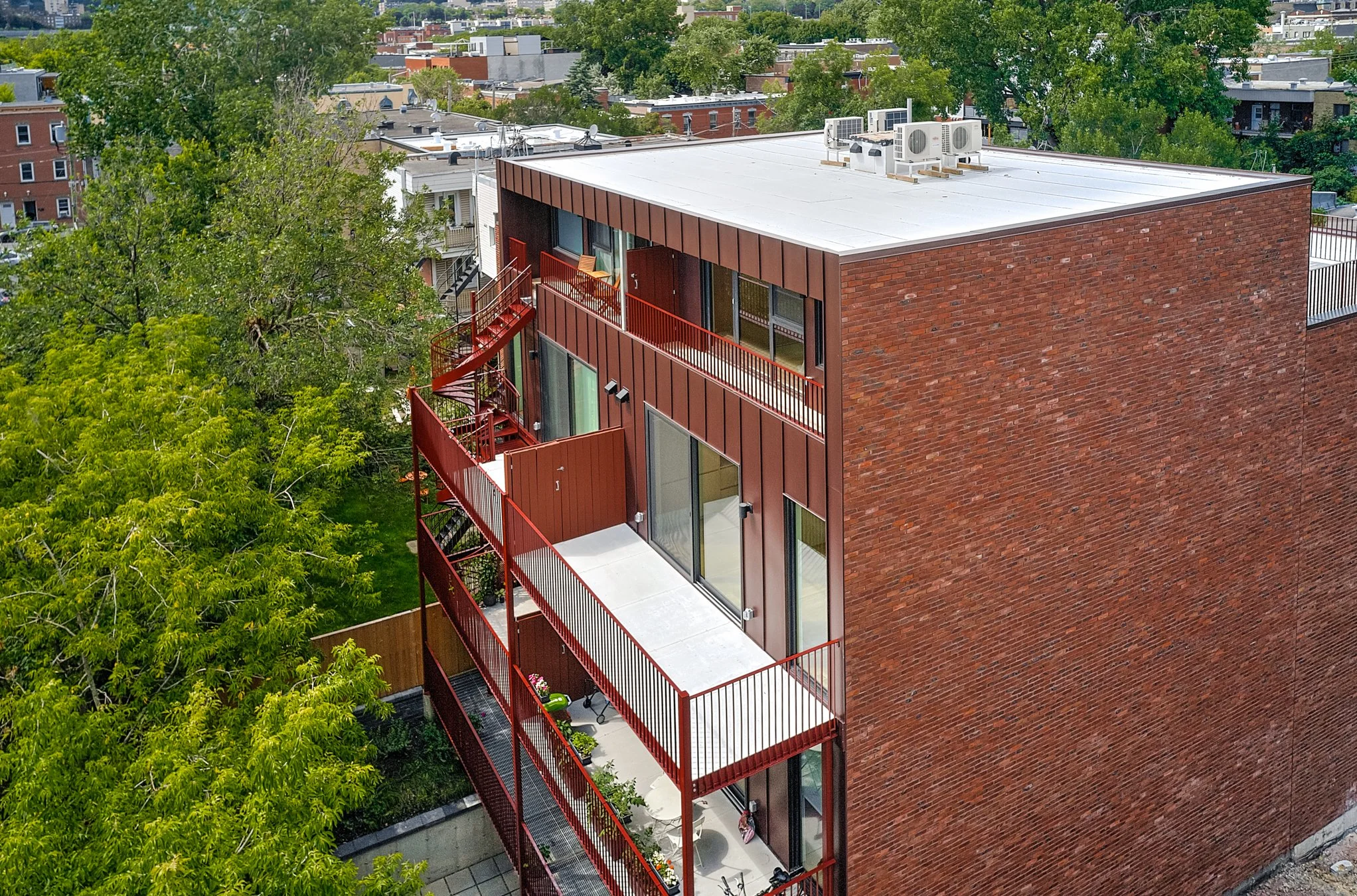 A multi-story residential building with red metal balconies on each floor, large glass sliding doors, and a brick side wall. The building is surrounded by trees and other residential structures in the background.