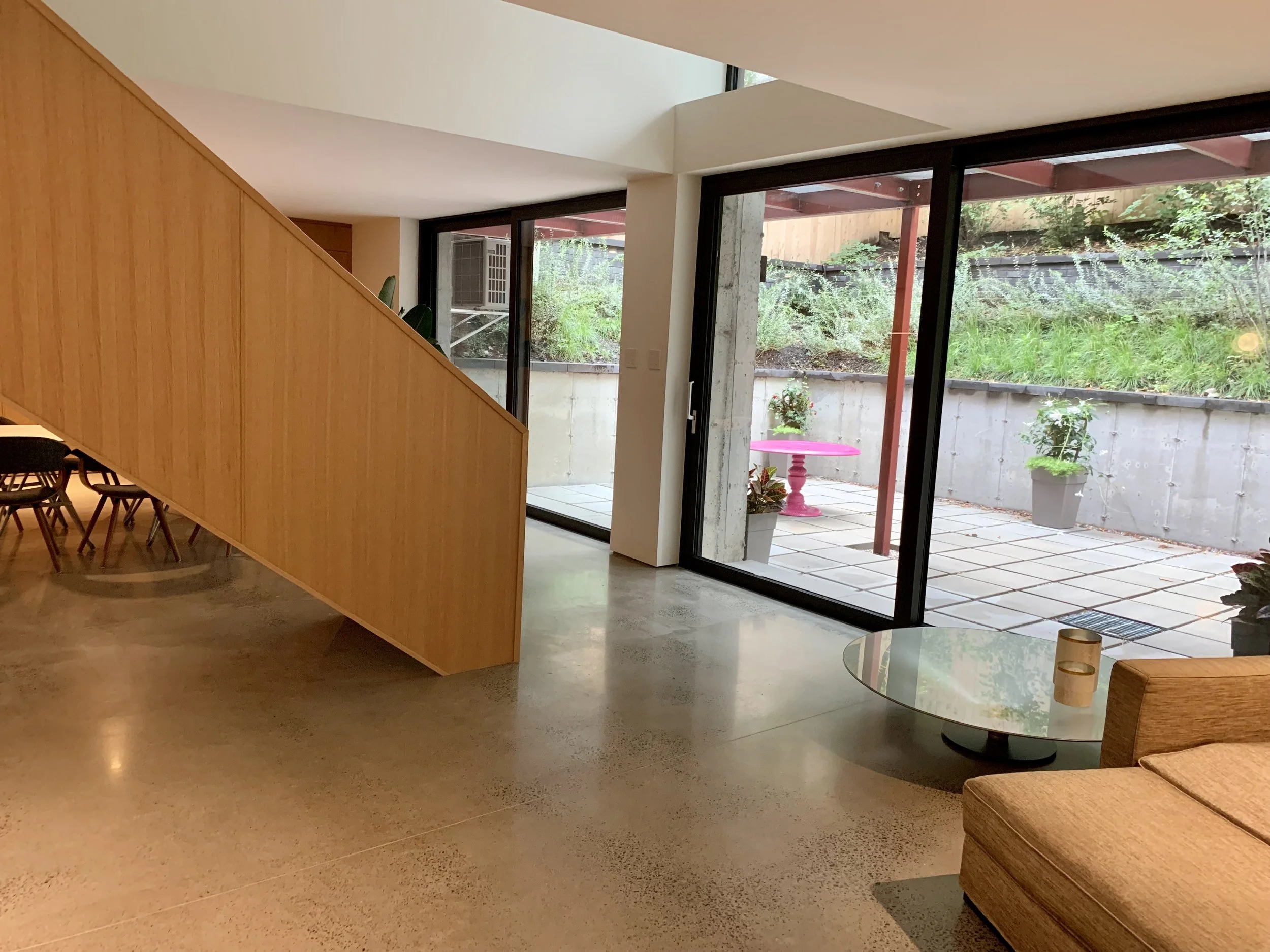 Interior view of a modern living space with a diagonal wooden partition, glass sliding doors leading to an outdoor patio, a beige couch, a round glass coffee table with a decorative candle, and greenery outside.