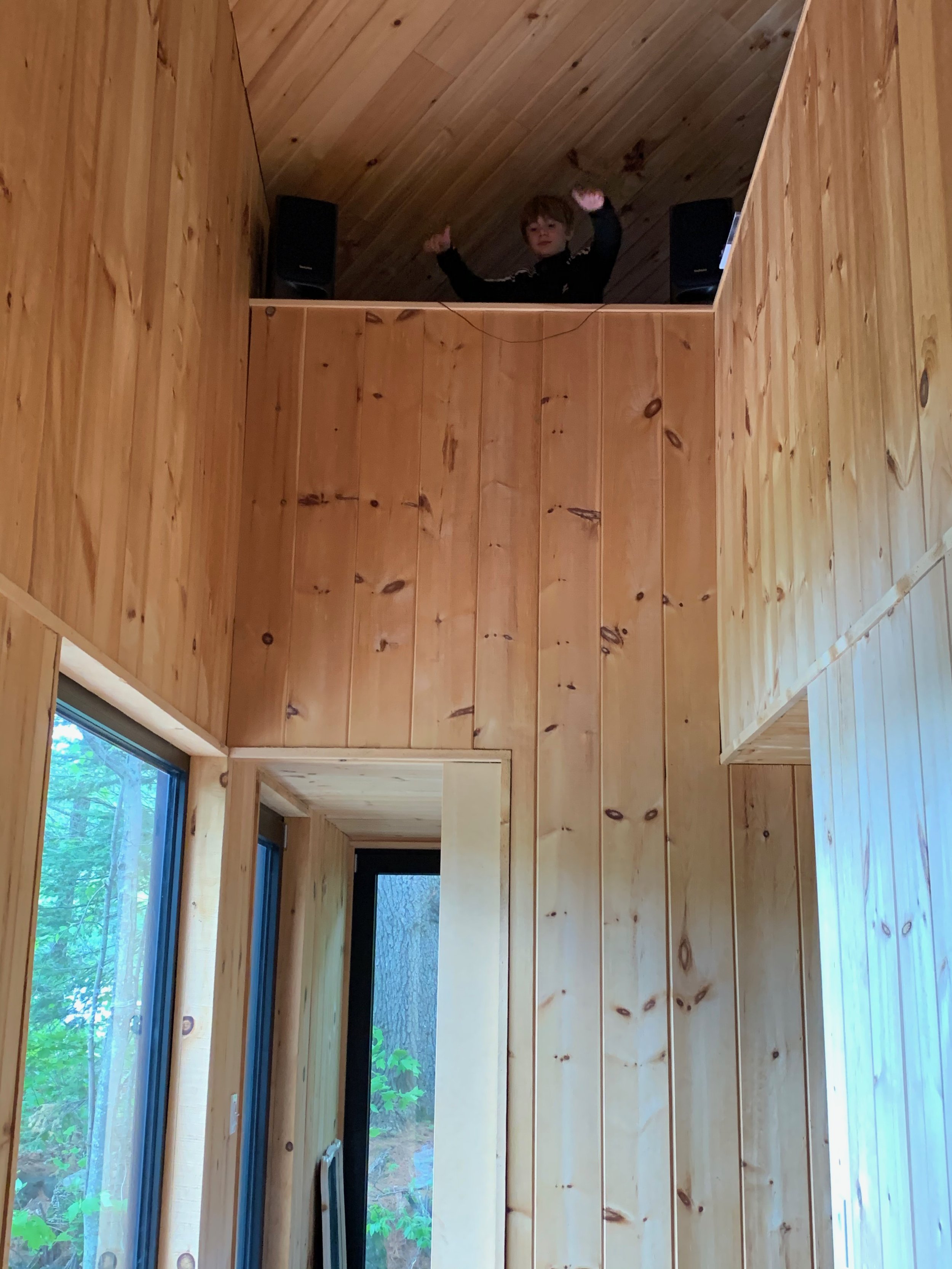 A person standing in an upper loft of a wooden interior, looking down. The room features natural wood paneling and large glass windows.