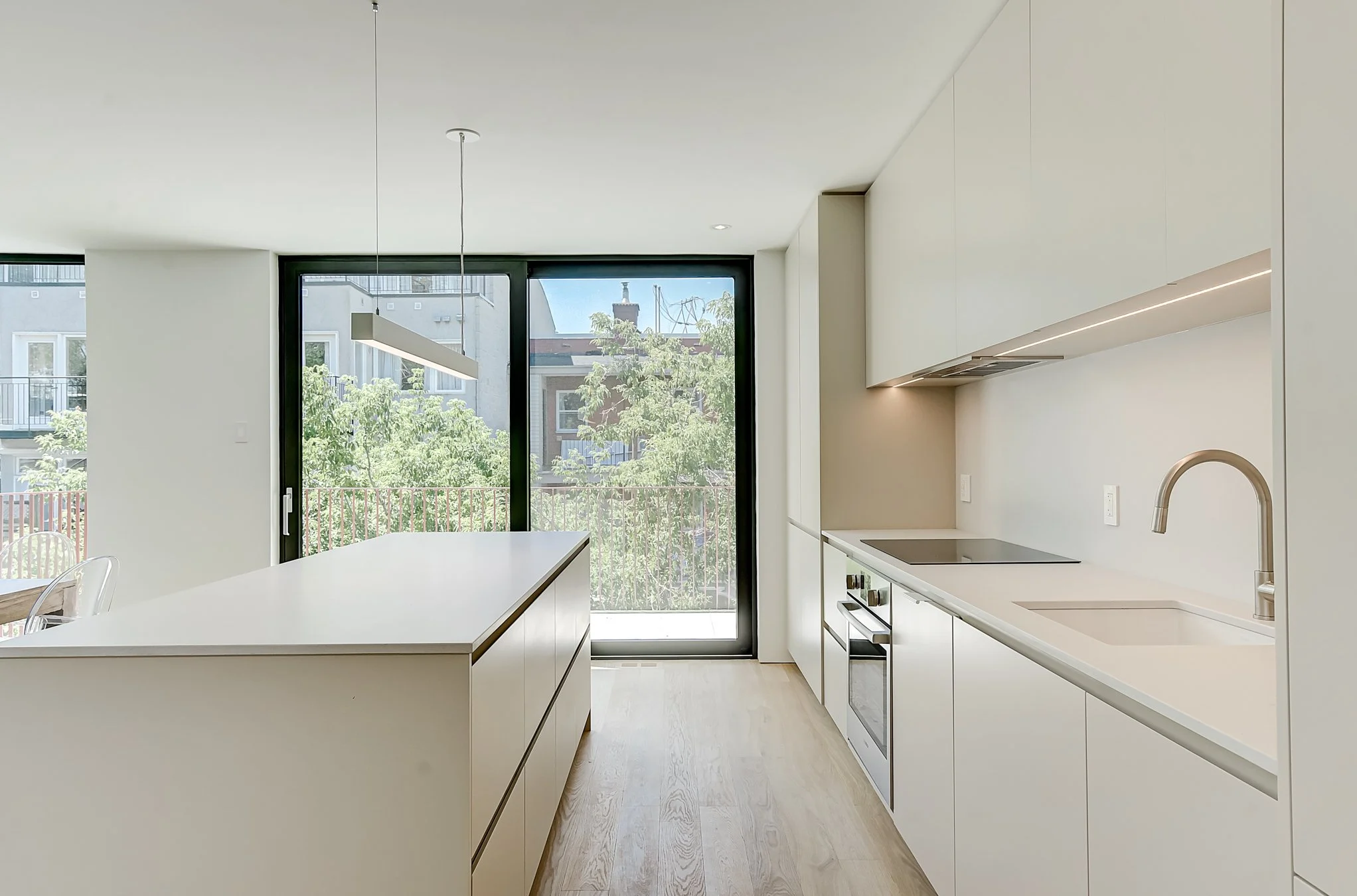 Modern kitchen with white cabinets, a large white island, a black sliding glass door leading to a balcony, and a view of neighboring buildings and trees outside.