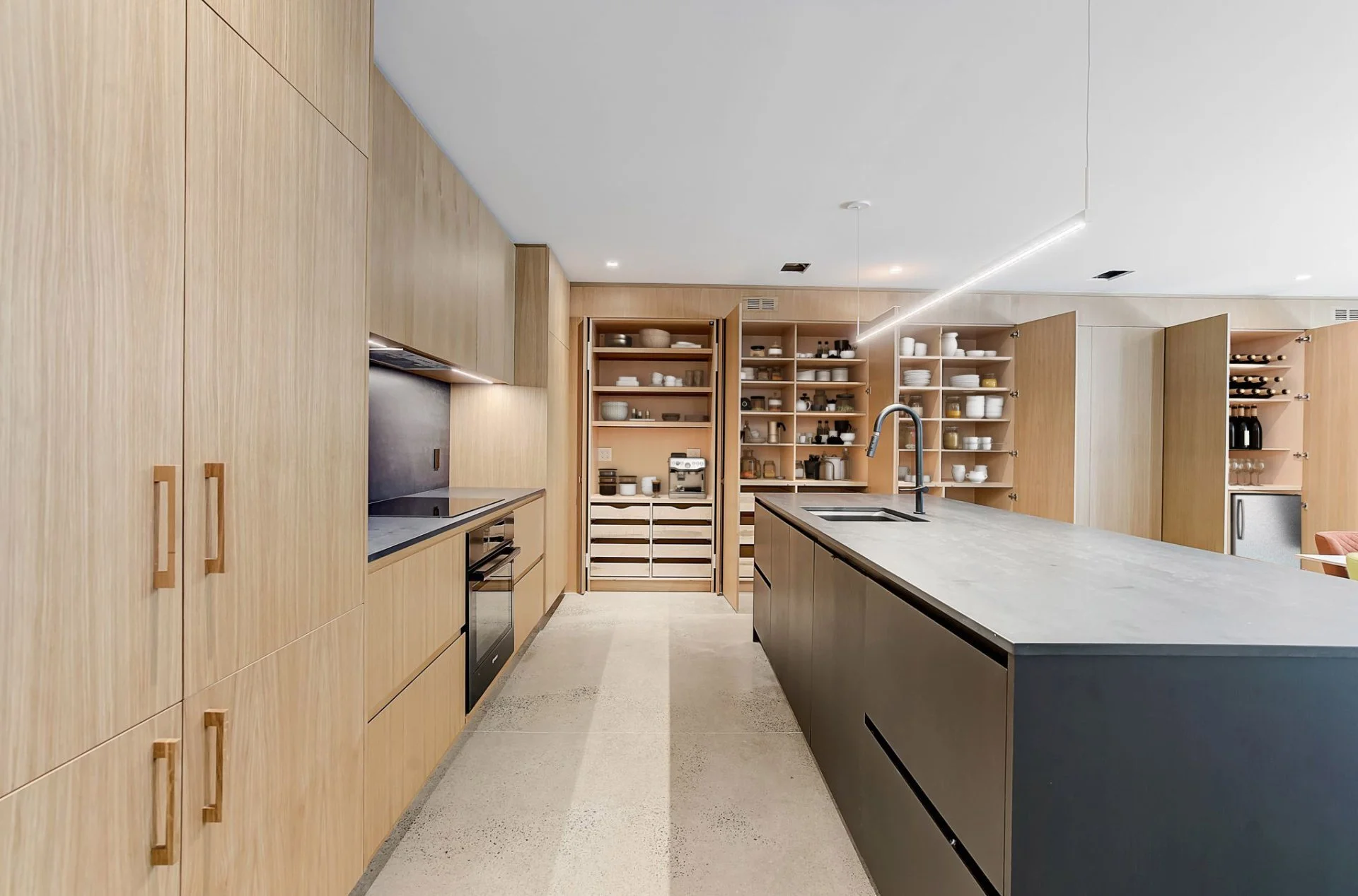Modern kitchen with light wood cabinetry, a gray island with a black faucet, open shelves with dishes and jars, and a concrete floor.