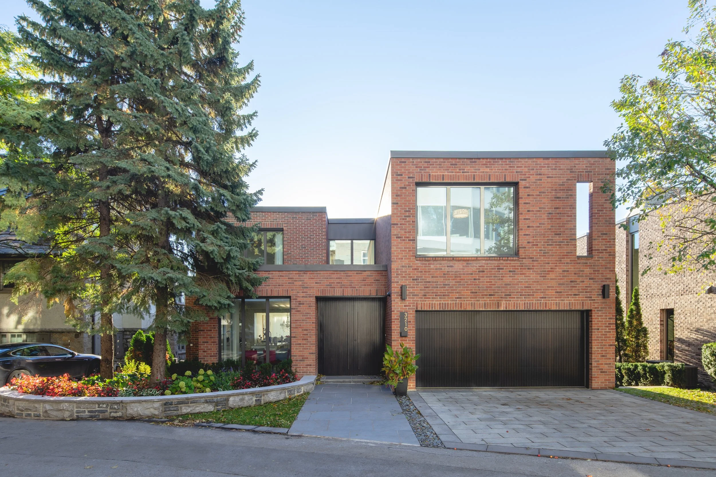 Modern brick house with large windows, attached garage, and landscaped yard with trees and flowering plants.