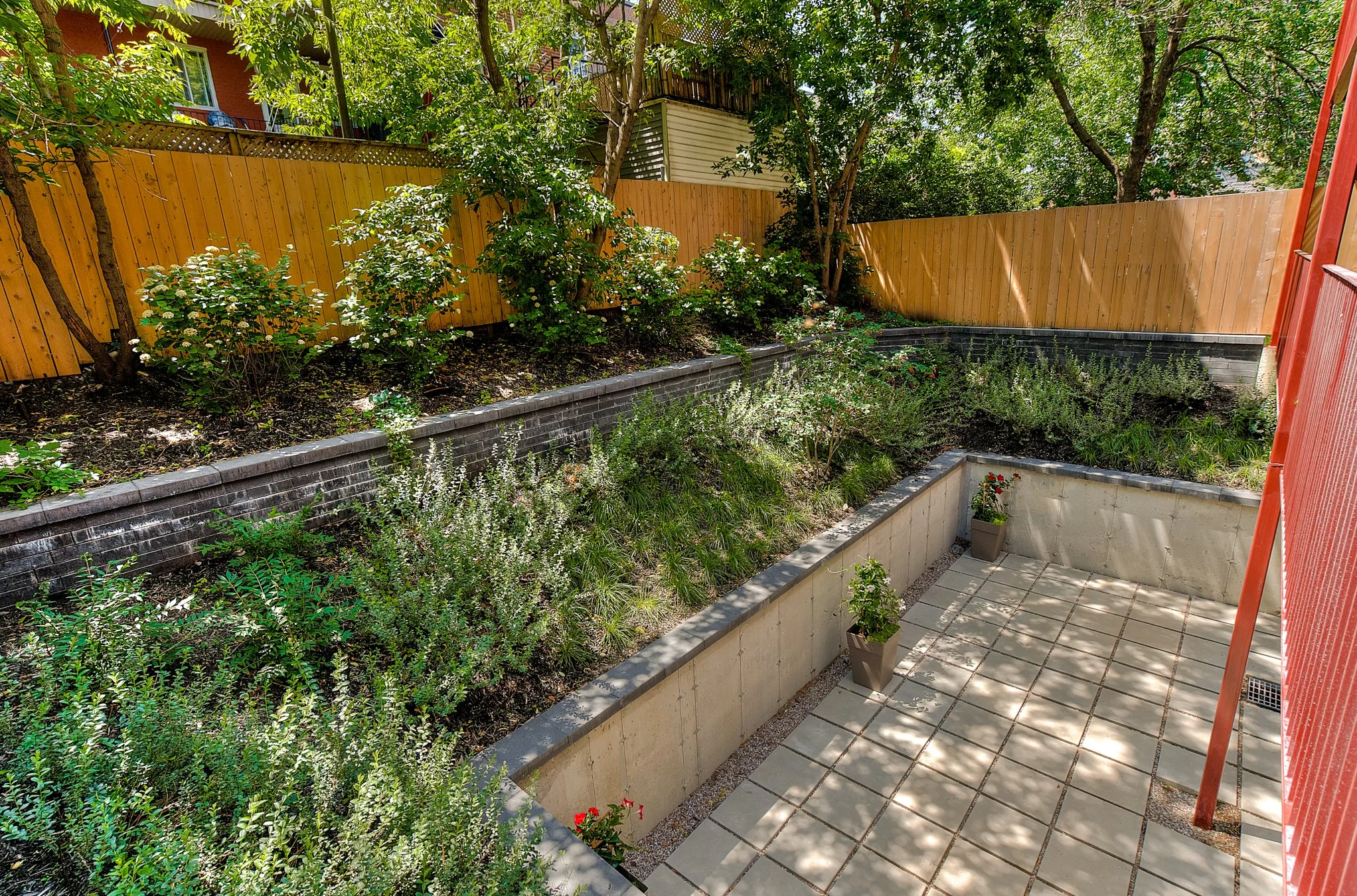 A backyard with a small tiled patio area with potted plants, surrounded by a flower bed with bushes and trees, enclosed by a wooden fence.