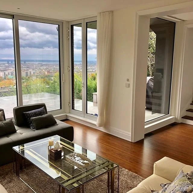 Interior of a living room with large glass doors leading outside, showing a city view. The room has a dark sofa with pillows, a glass coffee table, and an area rug, with natural light coming in.