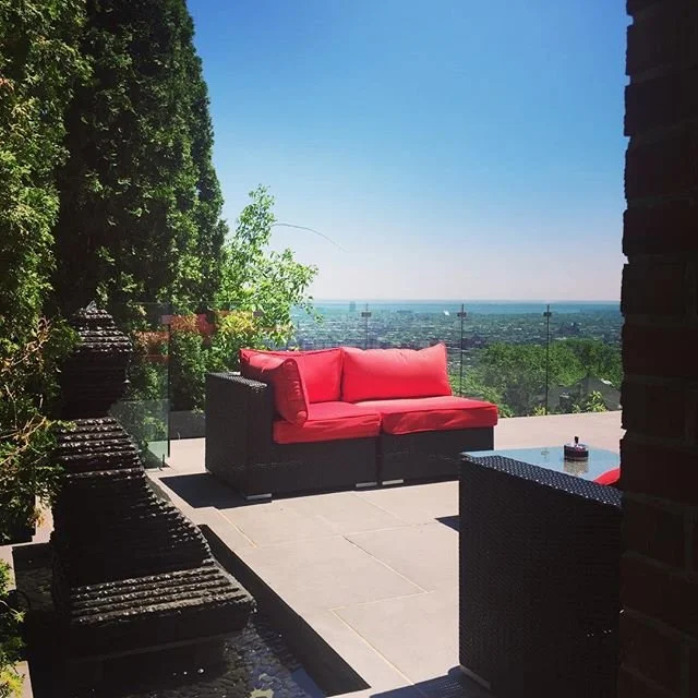 Outdoor patio with a red cushioned sofa, green trees, glass railing, and a city view under a clear blue sky.