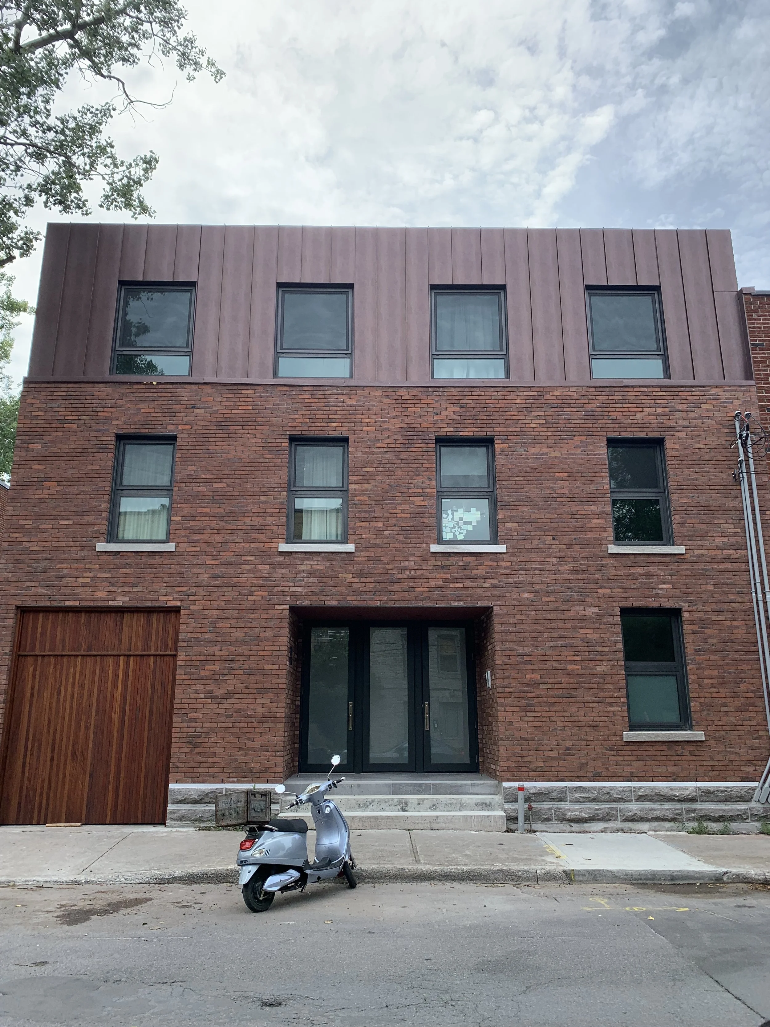 A modern brick building with a wooden garage door and a scooter parked in front, under a partly cloudy sky.