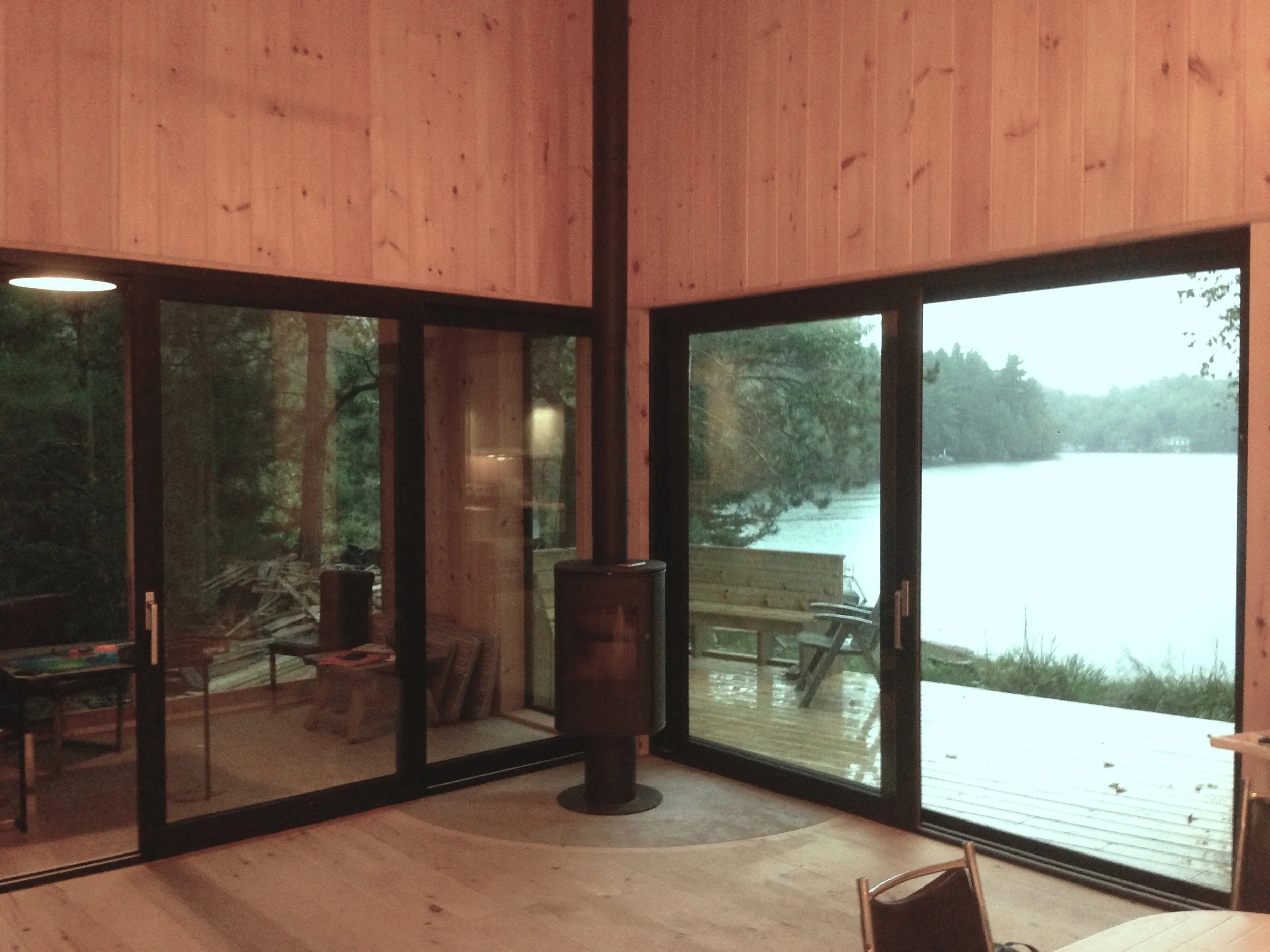 Interior of a wooden cabin with large glass sliding doors showing a lakeside view and a small wooden deck with outdoor furniture.