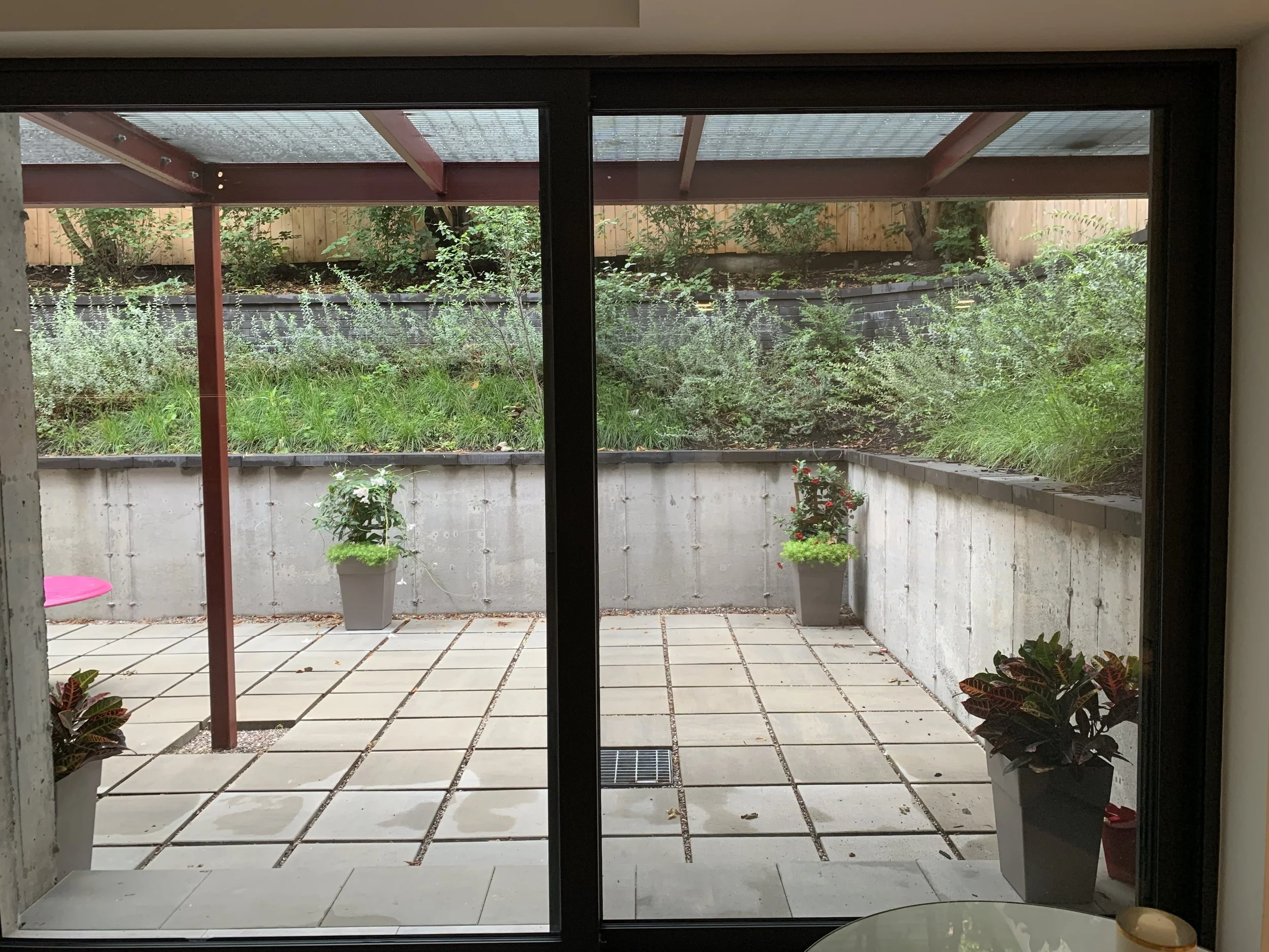 View of an outdoor patio through glass sliding doors, featuring potted plants, tiled flooring, and a garden with shrubs and a wooden fence in the background.