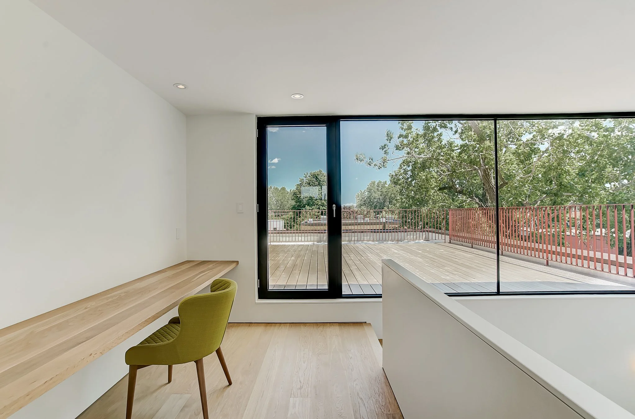 Minimalist room with large glass sliding doors leading to a balcony with a wood deck and a red railing, overlooking trees and a clear sky.