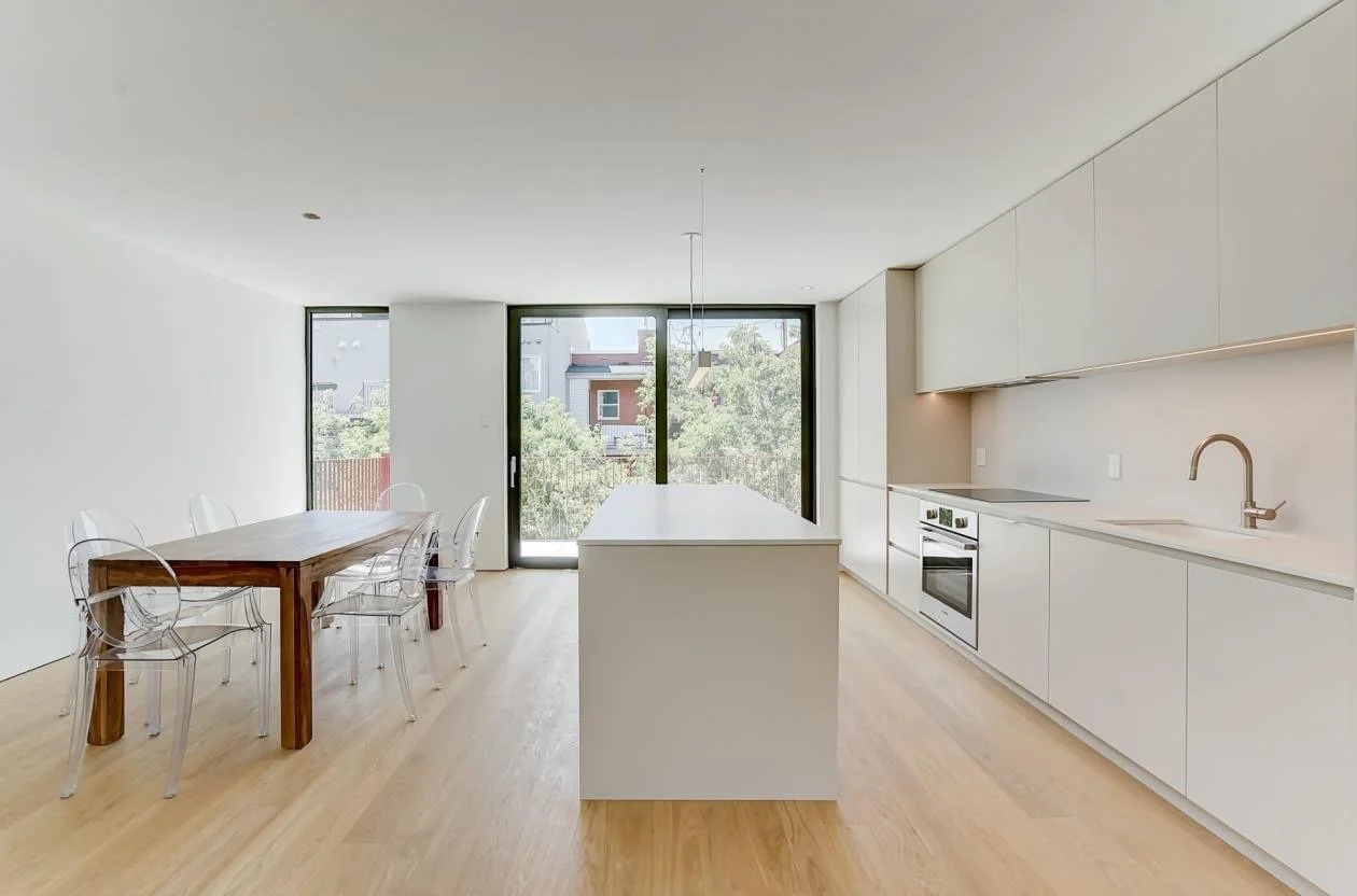 Modern kitchen with white cabinetry, island, and minimalist decor. Adjacent dining area with wooden table and transparent chairs, large glass sliding door opening to outside, natural light, light wood flooring.