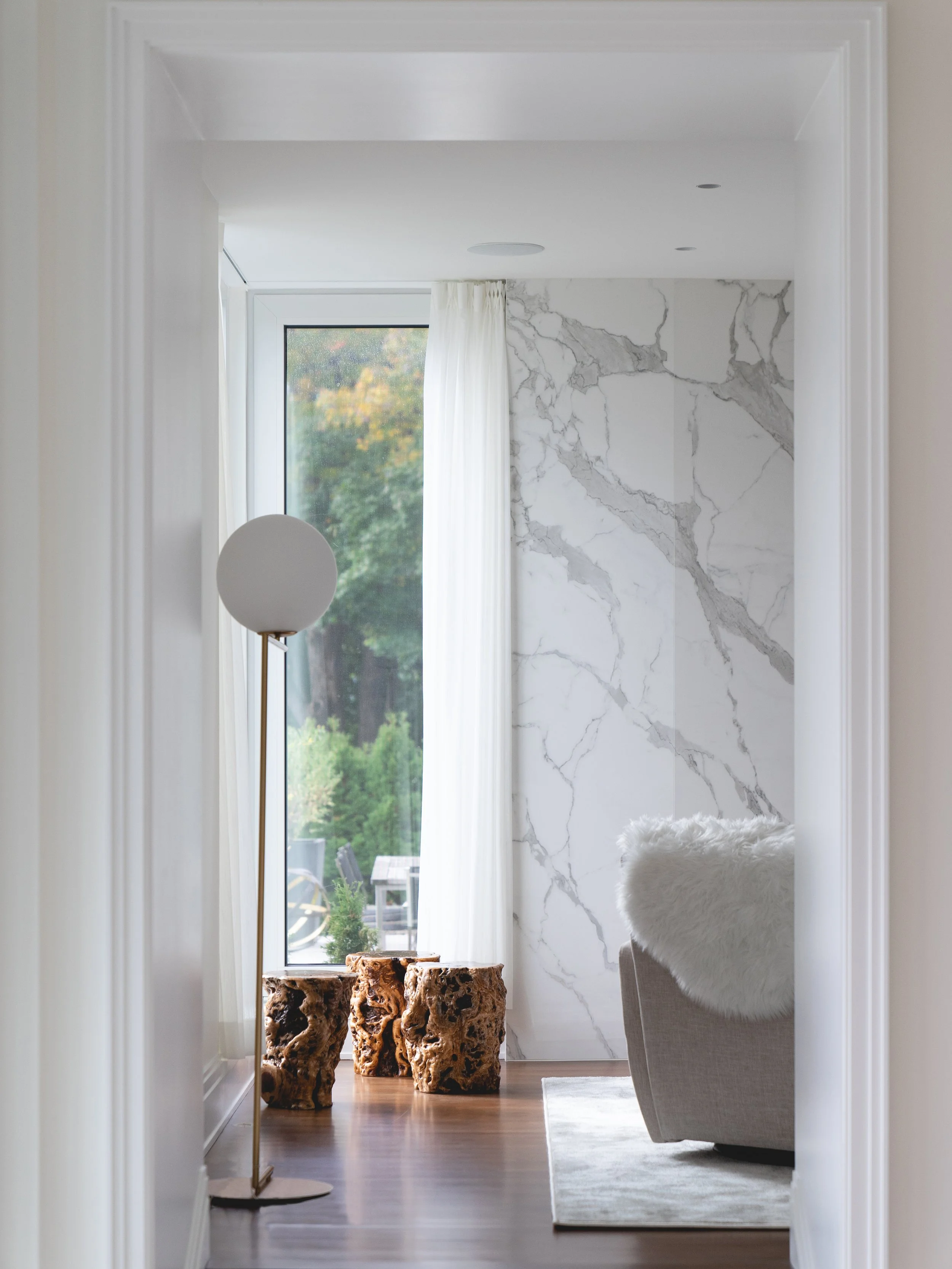 A modern living room with a large window, white curtains, a marble accent wall, a beige sofa with a furry blanket, three textured wooden stools, a white floor lamp, and a view of greenery outside.
