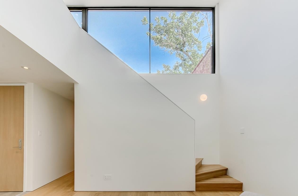 Interior of a modern, minimalistic house with a staircase, wooden stairs, white walls, a large window showing trees and a blue sky.
