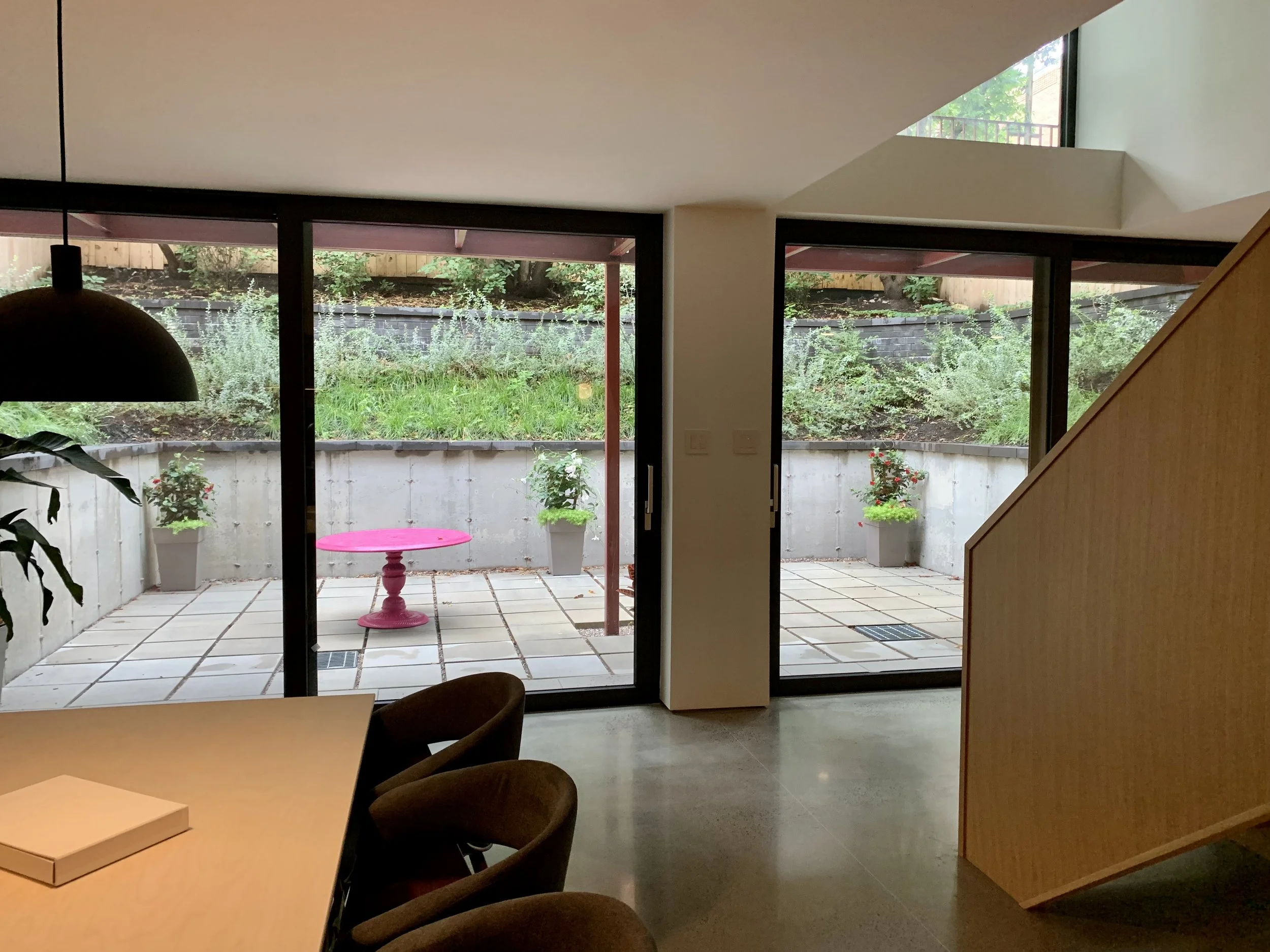 Indoor view of a room with a large window and sliding glass door leading to a small patio with potted plants and a pink table.