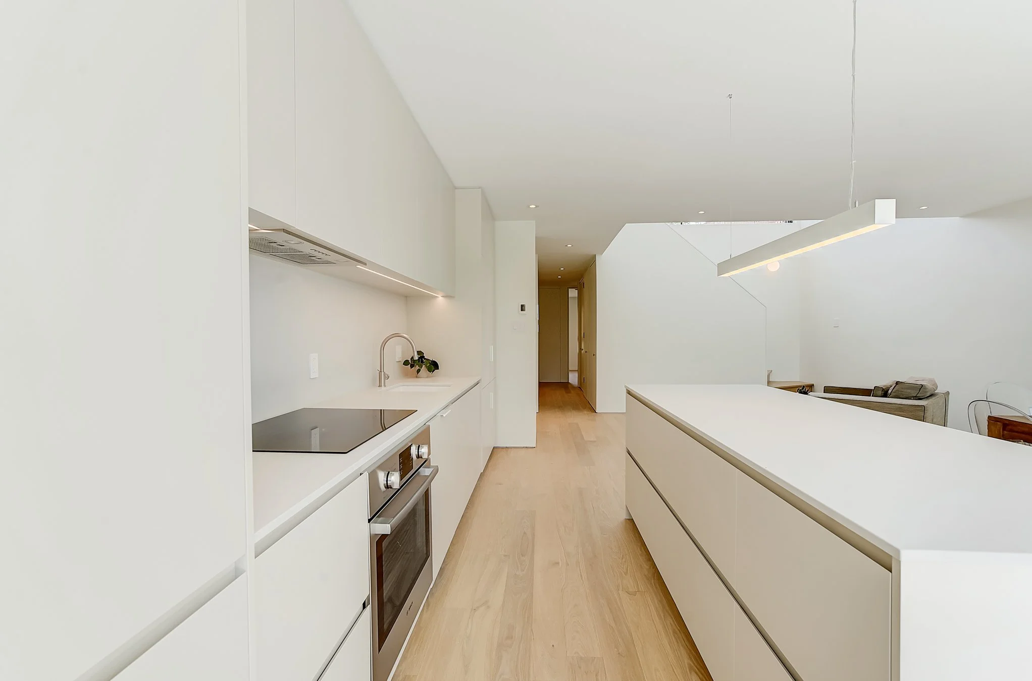 Modern minimalist kitchen with white cabinetry, wooden flooring, and a kitchen island, featuring built-in appliances and a hanging light fixture.