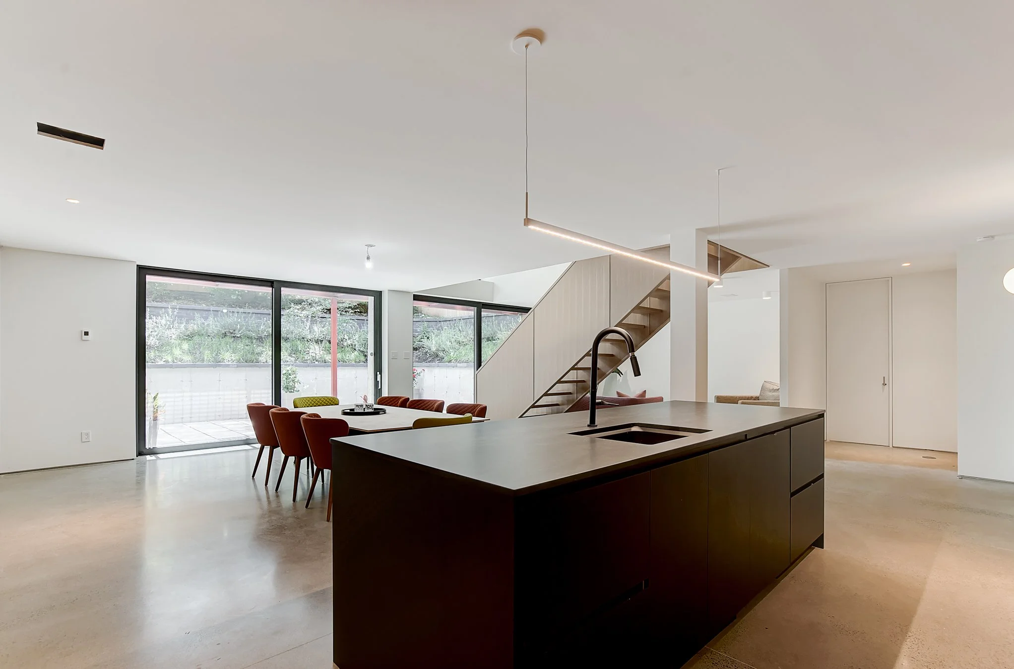 Modern open-concept kitchen and dining area with large sliding glass doors, a black kitchen island with a sink, colorful dining chairs, and a staircase leading to the upper level.