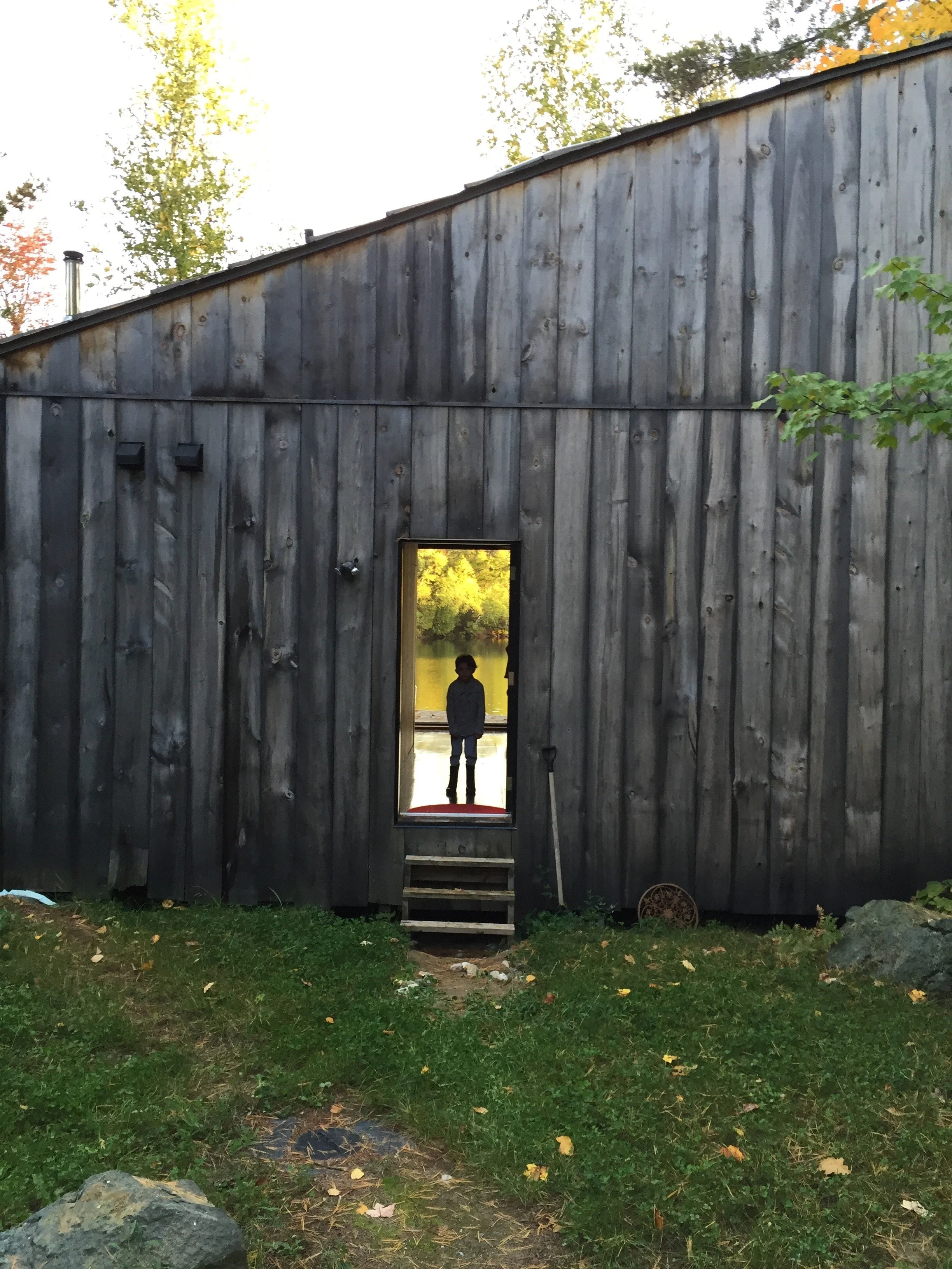 Person standing inside a small barn with a rectangular mirror reflecting a lake and trees outside.