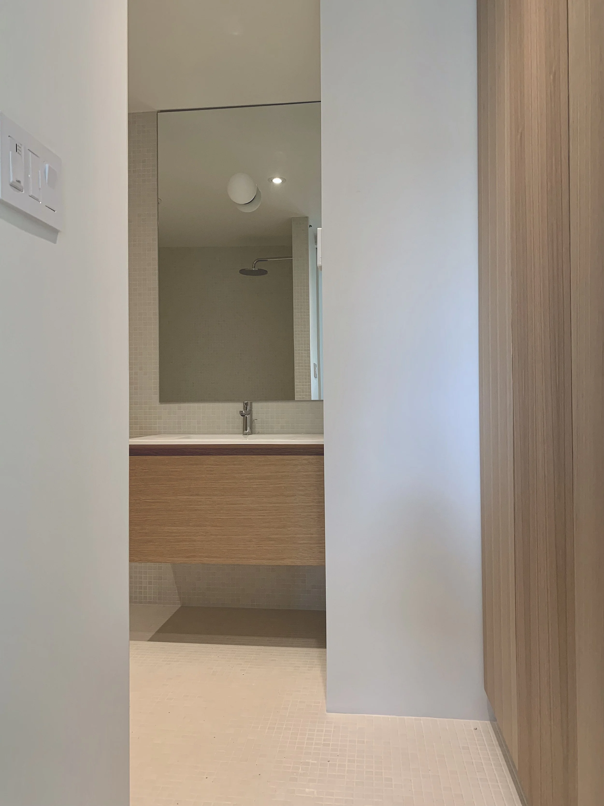 Modern bathroom with mirror, wooden vanity, white sink, beige tiled floor, and shower with overhead rain shower, seen through a doorway.