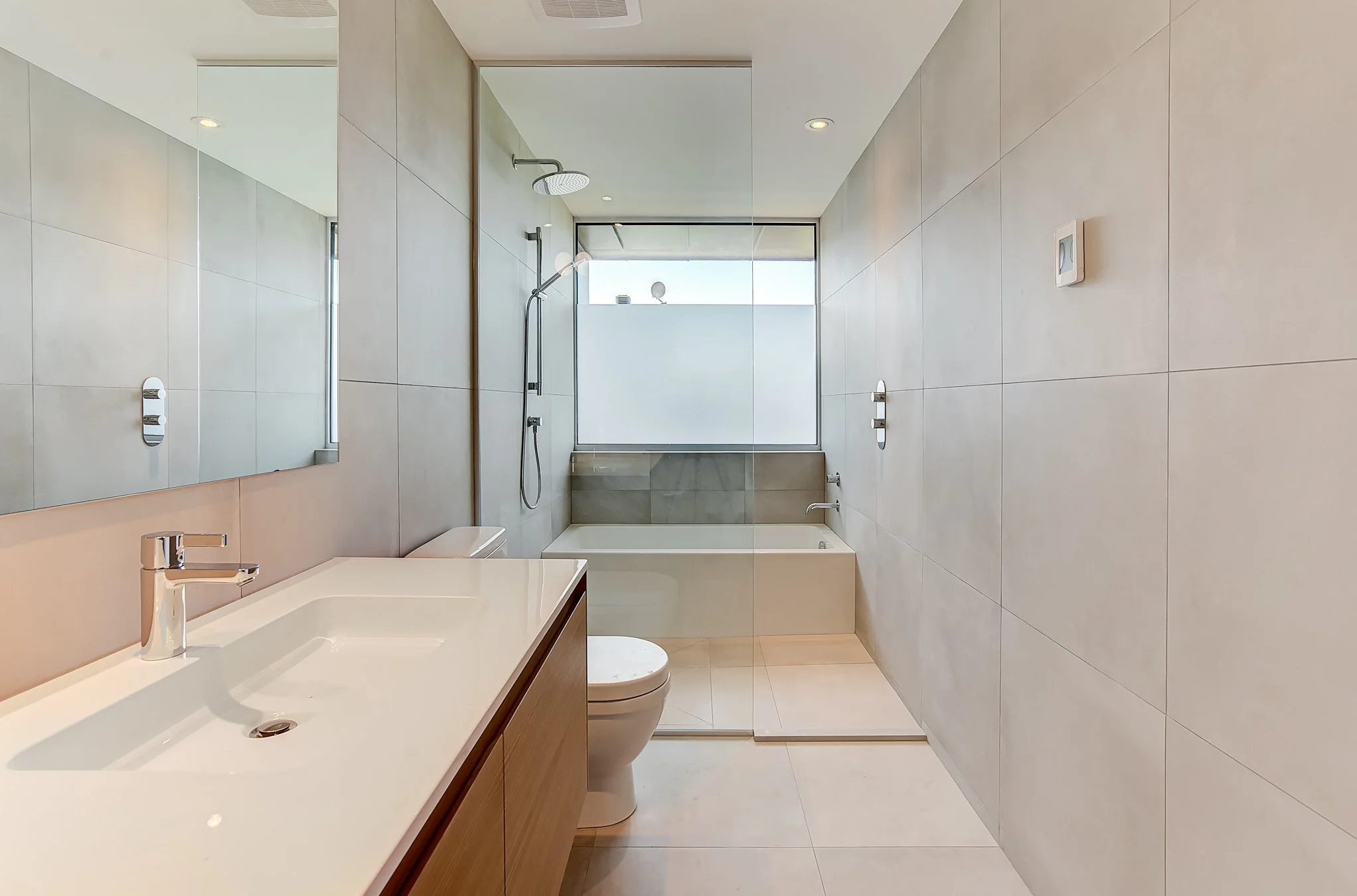 Modern bathroom with a large mirror, beige wall tiles, a white sink with a wooden cabinet, a toilet, a glass-enclosed shower area, a bathtub near a frosted window, and recessed ceiling lights.