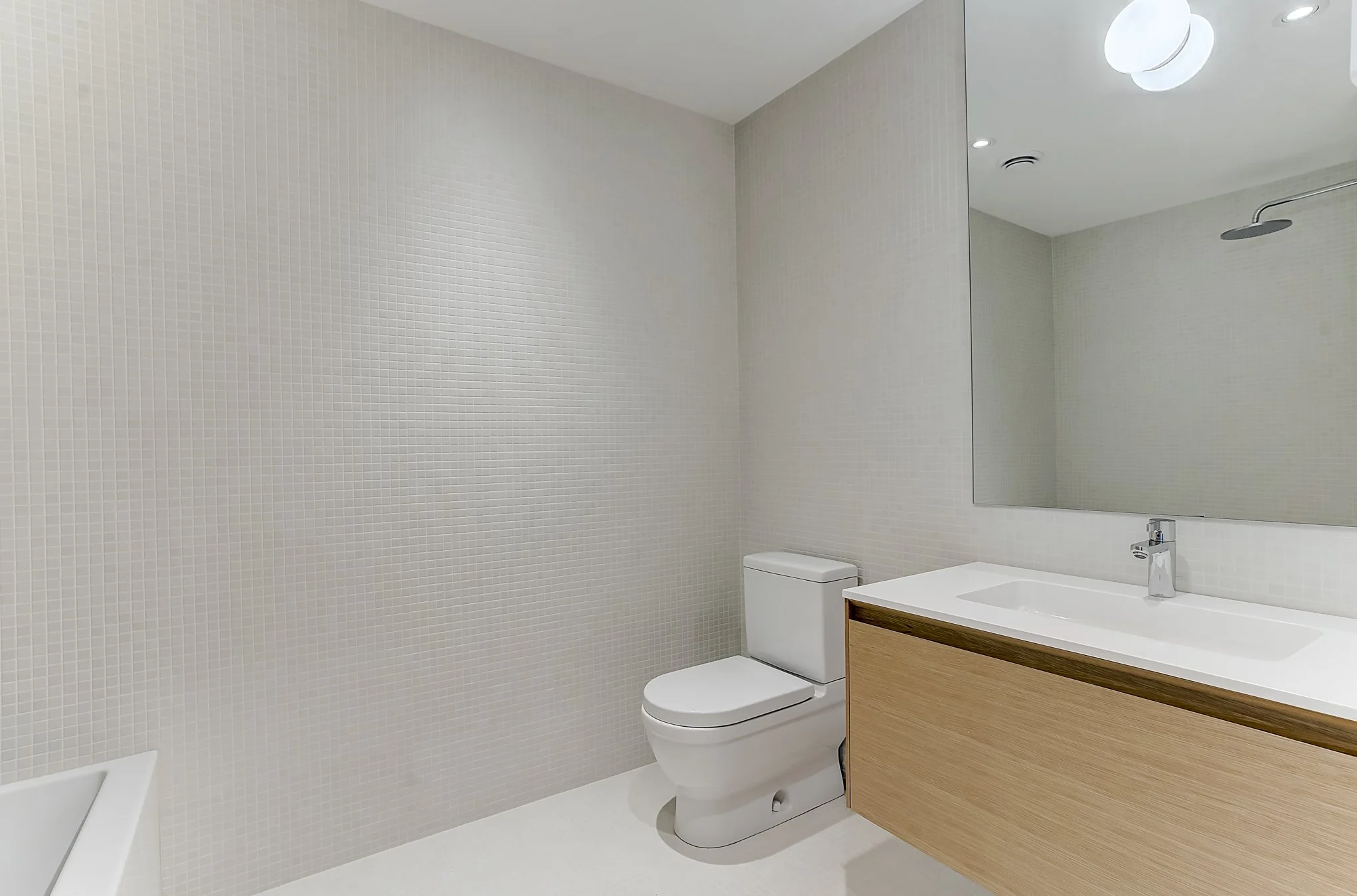 Modern bathroom with a white toilet, a wooden vanity with a white sink, a large mirror, and a walk-in shower with a ceiling-mounted showerhead. The walls are tiled with small white tiles.