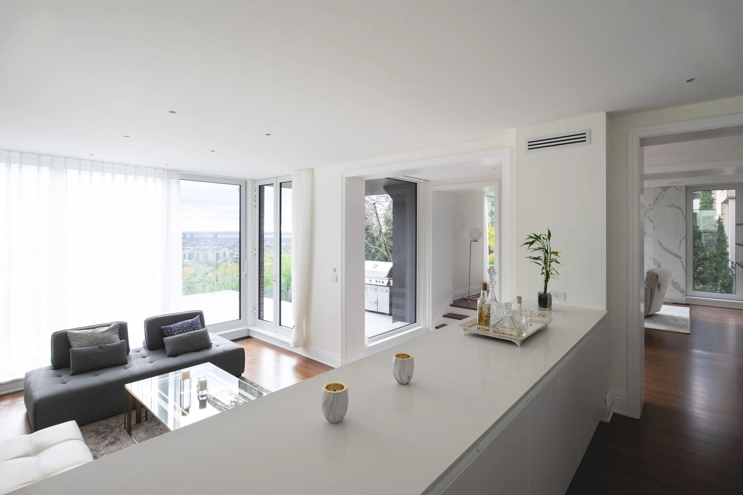 Modern living room with large floor-to-ceiling windows, gray sofa, glass coffee table, and white walls, overlooking a city view.