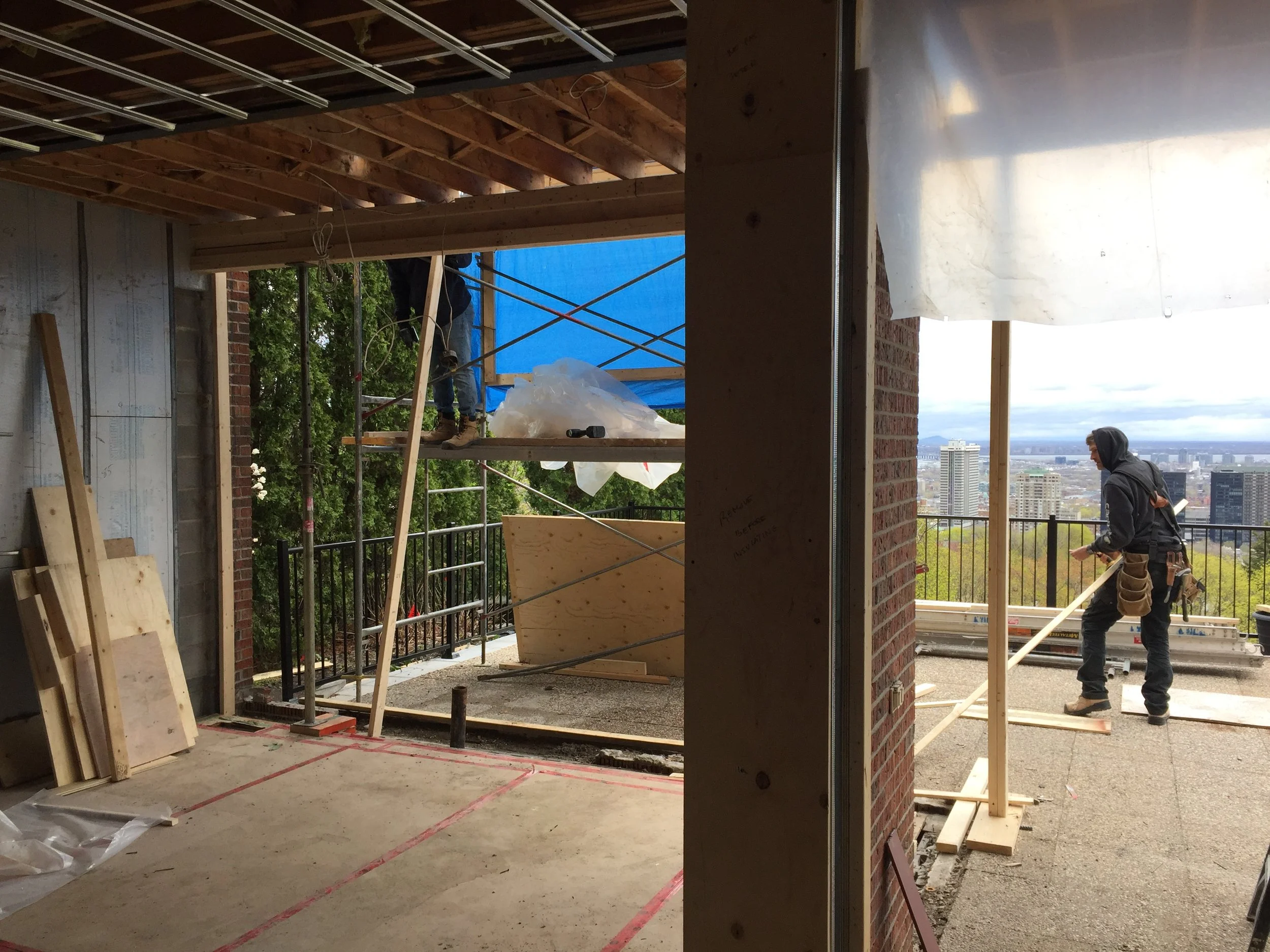 Construction workers are building a balcony on a high-rise building. One worker on the right is pushing a wooden plank, and another worker is on scaffolding outside. The view shows a city skyline with skyscrapers and green trees.