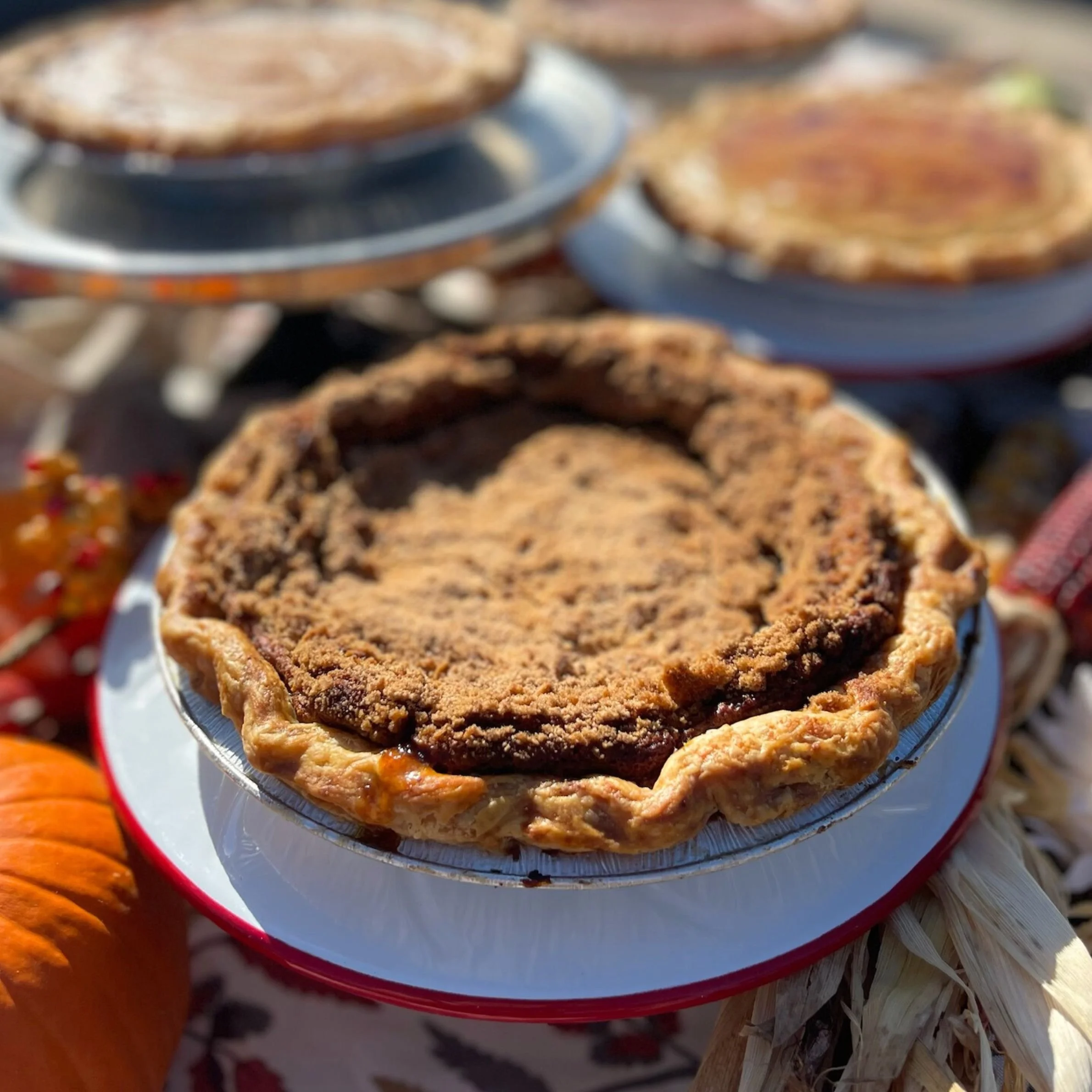 Close-up of a slice of pumpkin pie with a crumb topping on a red and white plate, surrounded by pumpkins and fall-themed decorations.