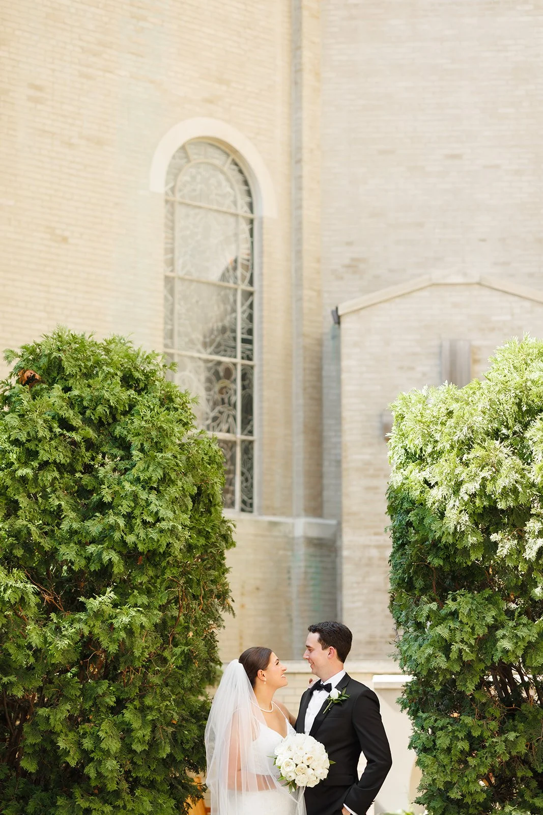 Outdoor wedding portrait in Texas of a smiling bride and groom standing between tall green trees with a historic church in the background, captured in golden hour light.