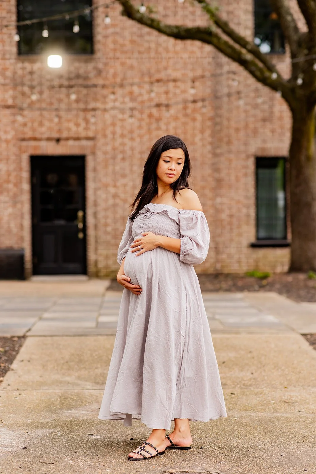 Pregnant woman in gray dress standing outdoors near brick building.