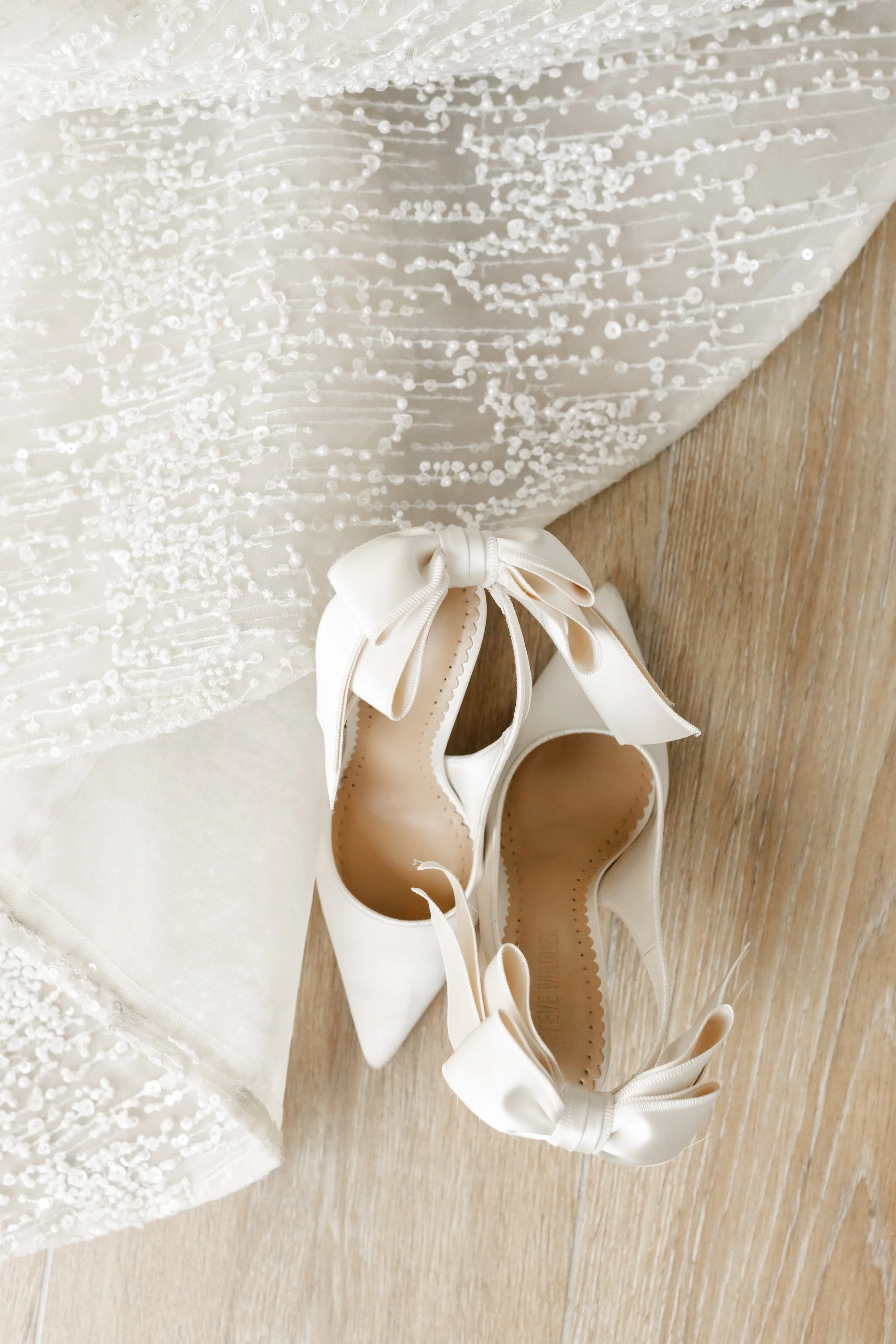 photographed by Wild Ivy Creative Photography, Pair of cream-colored satin high-heeled shoes with large bows on the back, placed on a wooden floor next to a white, beaded dress.