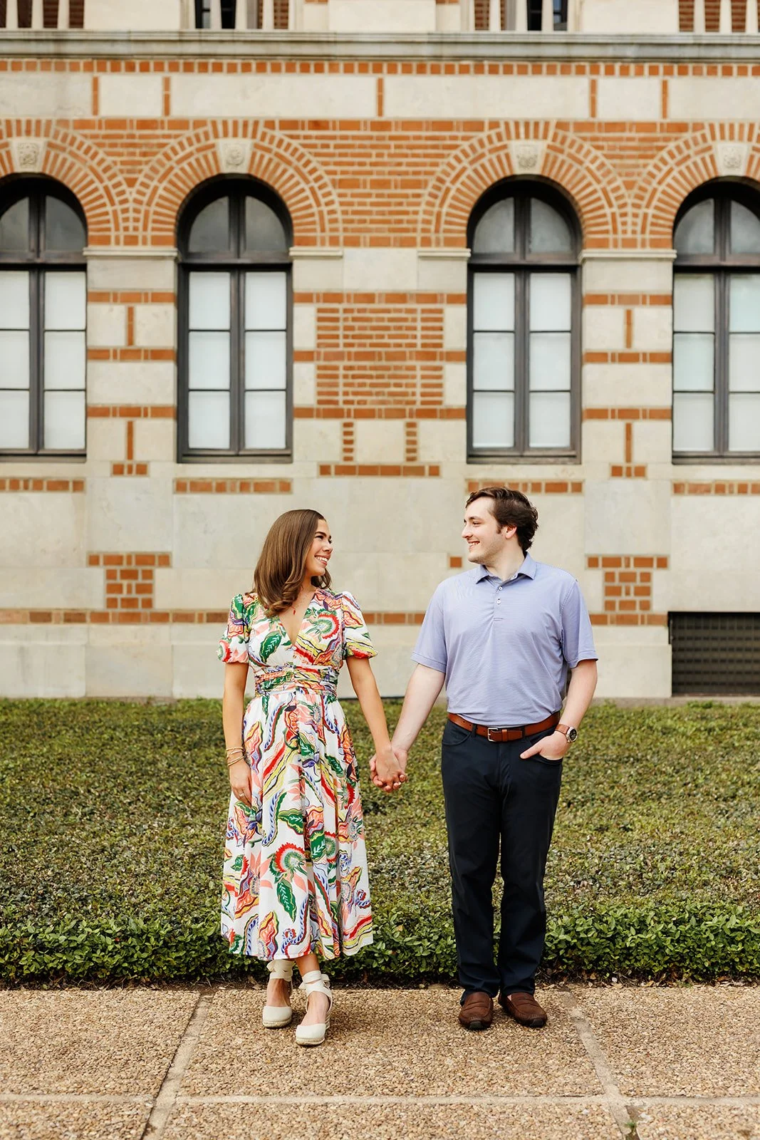 Engagement session with a happy couple walking in front of an ornate red brick building with arched windows in Houston, Texas.
