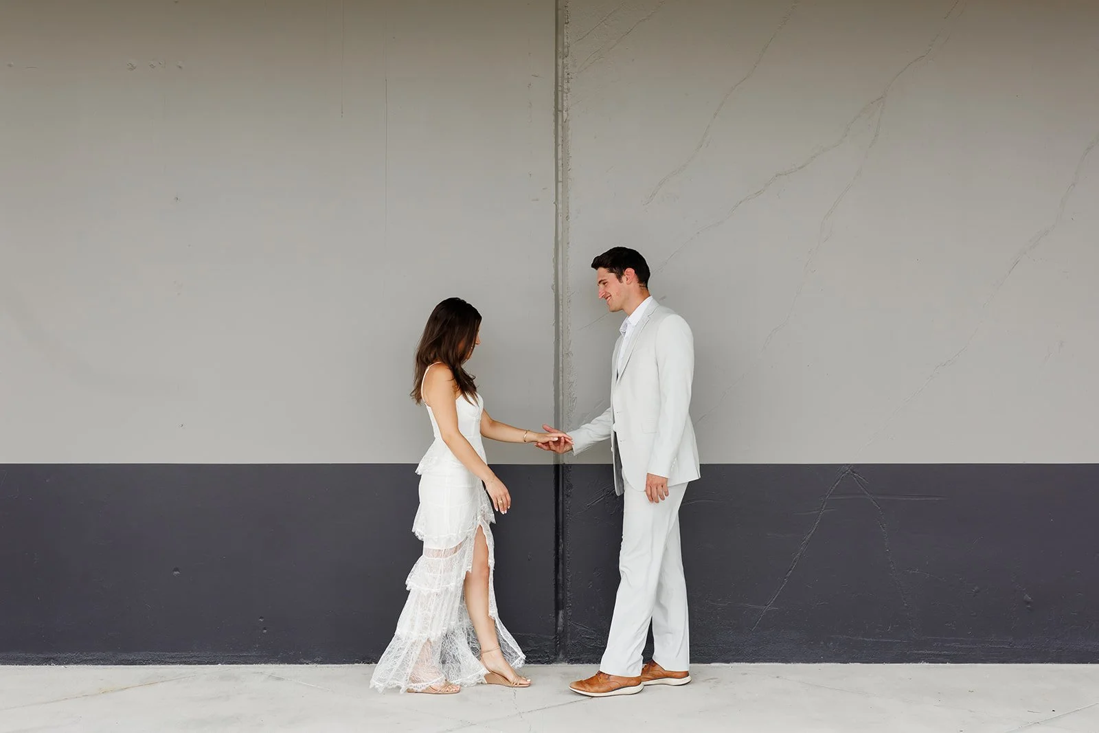 A couple in white attire holding hands in front of a large gray wall, standing close and smiling at each other.