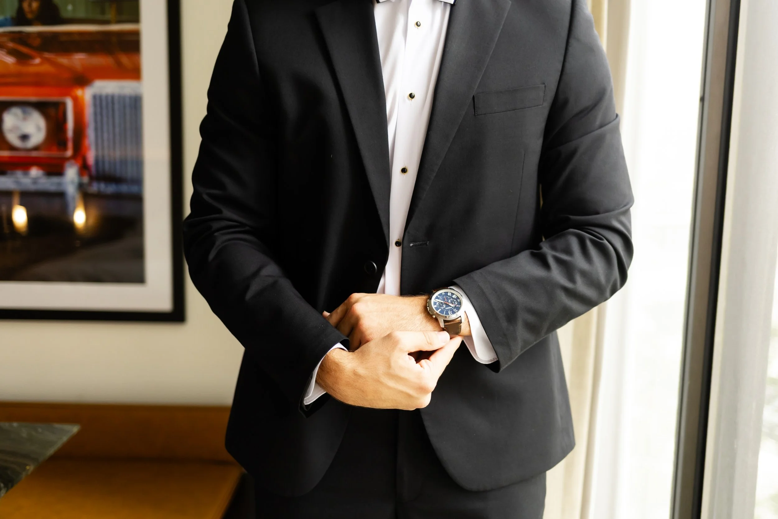 A man in a black suit and white shirt adjusting his watch in a room with framed pictures and a window with curtains captured by a woodlands area wedding photographer