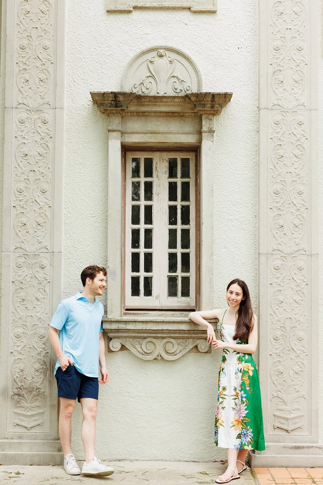 Outdoor engagement photo with couple standing near a grand arched window of a light stone building in the Heights of Houston photographed by Wild Ivy Creative Photography.
