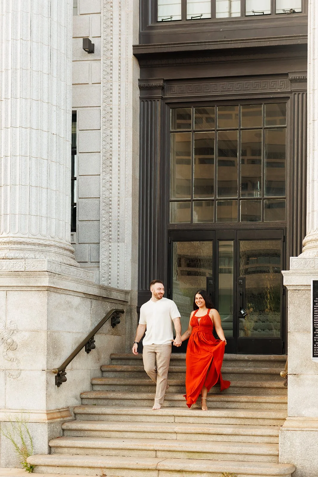 A couple holding hands and walking down the steps of a historic building with large columns, glass doors, and ornate architectural details.