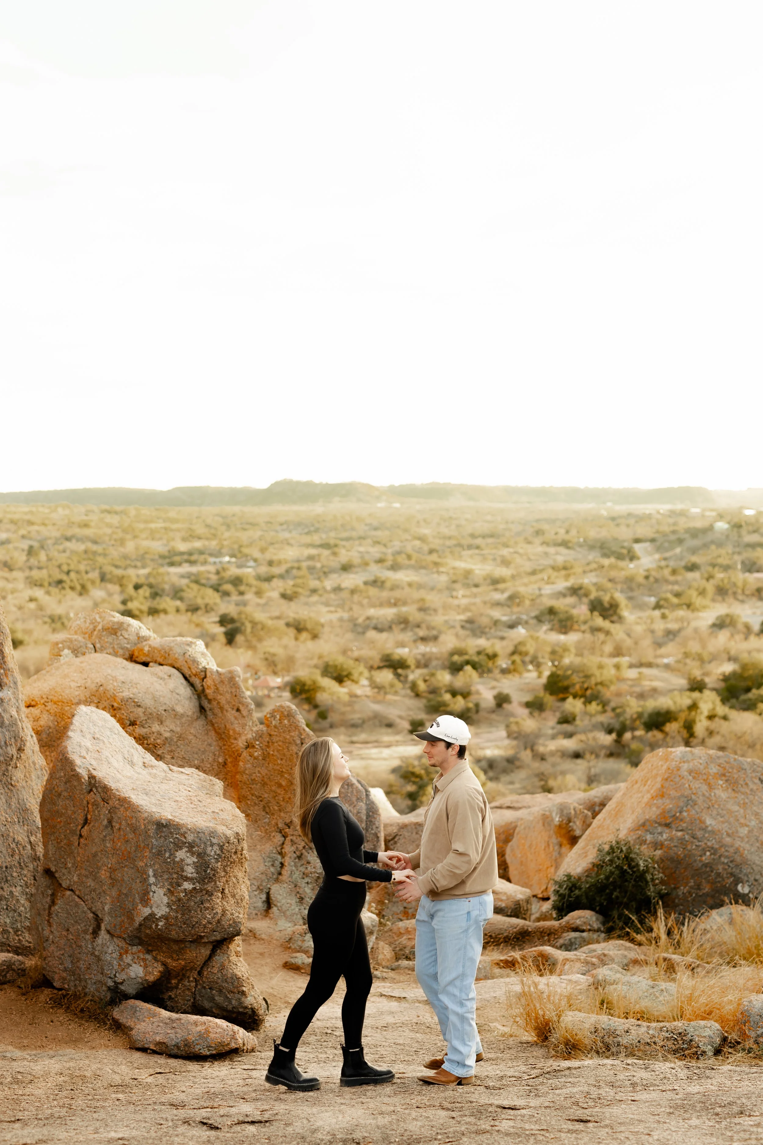 A man and woman holding hands and facing each other outdoors amongst large rocks, with a desert landscape and cloudy sky in the background.