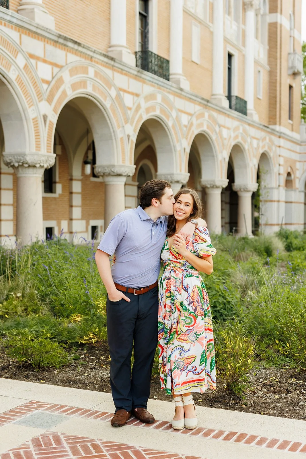 A couple standing outdoors near a building with arched brick and stone columns. The man is kissing the woman's cheek, and she is smiling. The woman is wearing a colorful patterned dress, and the man is dressed in a light blue shirt and dark pants.