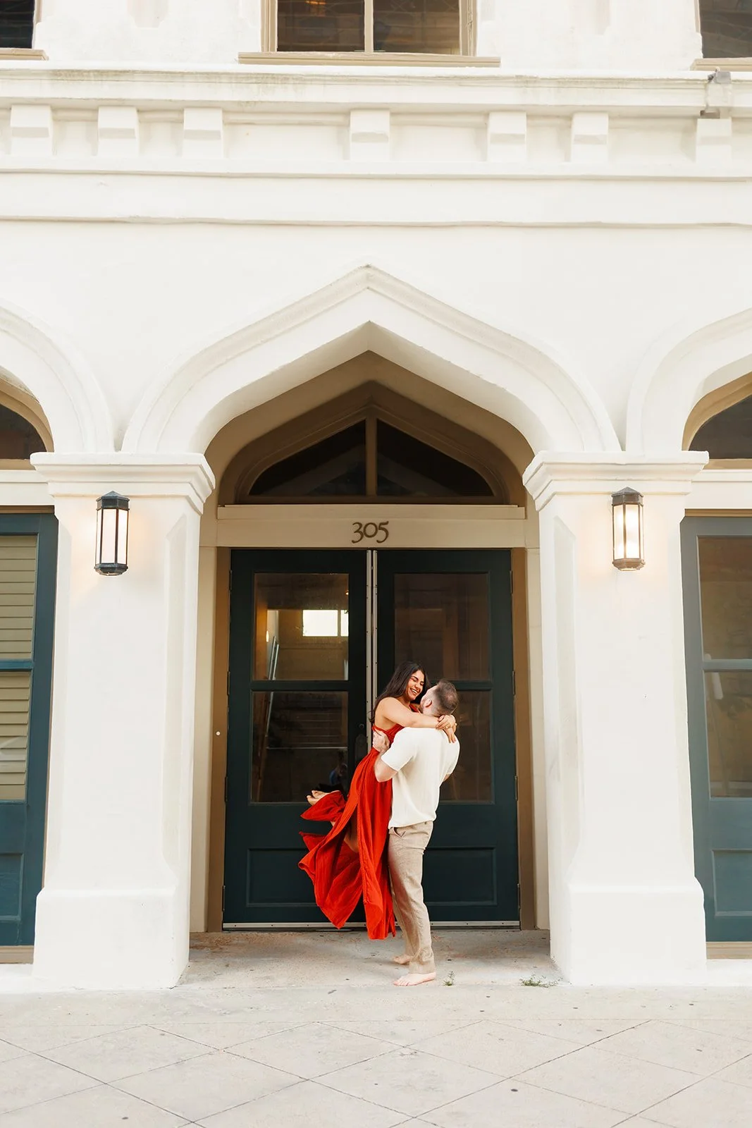 Playful and romantic Houston engagement photo in front of house number 305 in Galveston, TX, as the man lifts his fiancée in a red dress — photographed by Wild Ivy Creative.