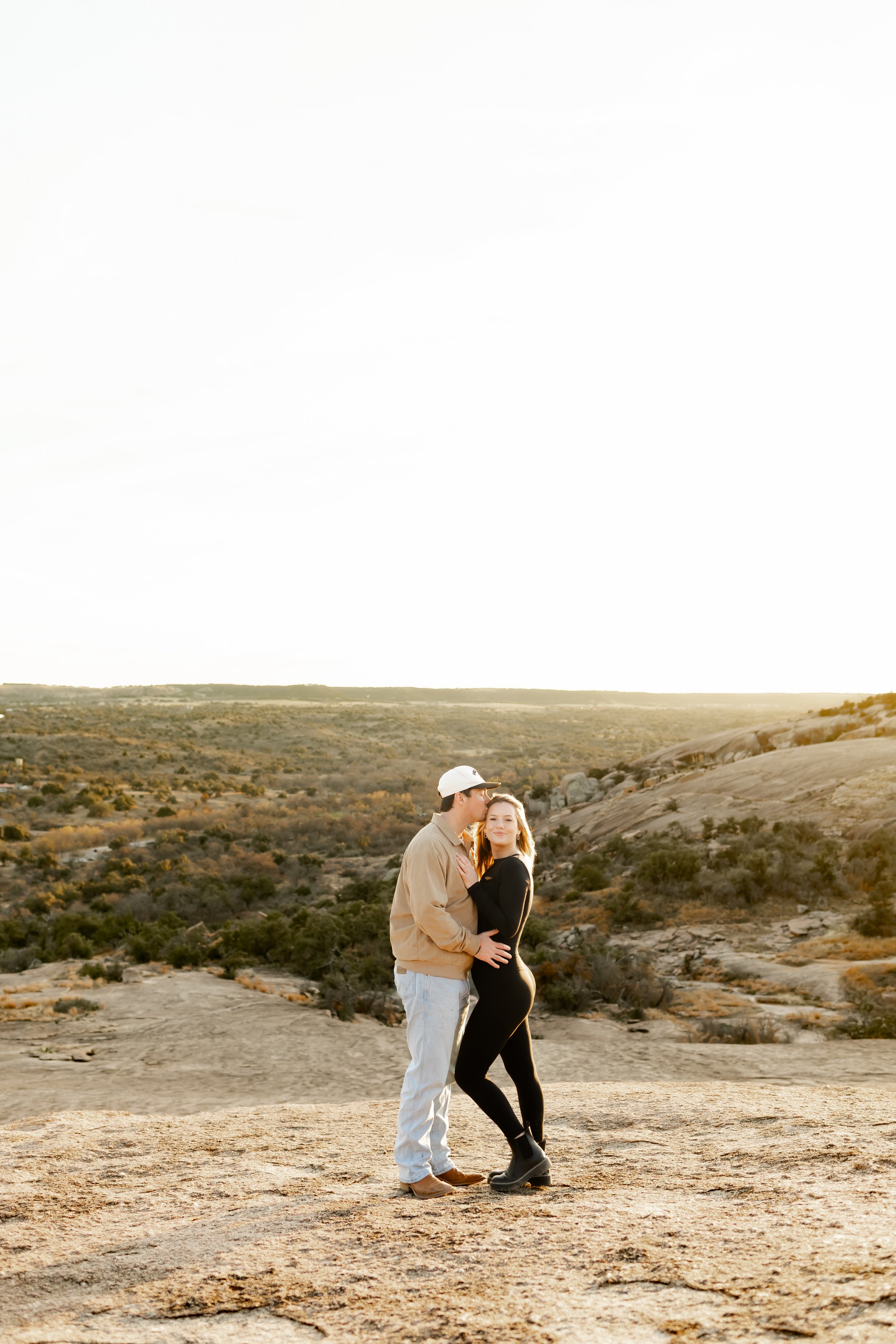 A couple stands closely together on a rocky desert landscape during sunset, with orange and yellow hues in the sky and sparse vegetation in the background.