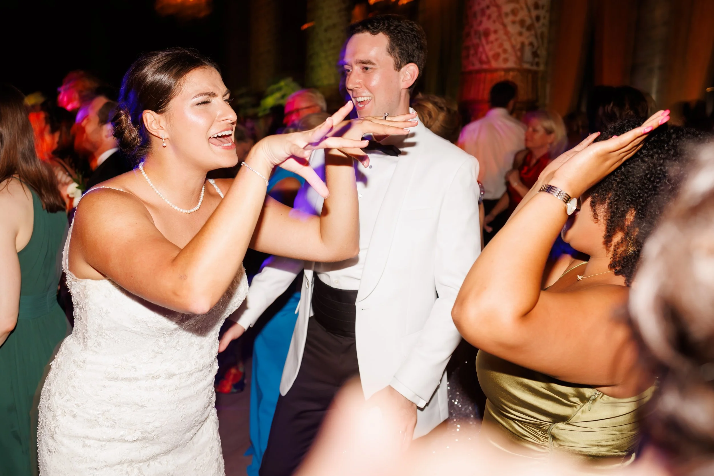 Bride in pearl necklace dancing at a Houston, Texas wedding reception, singing with joy beside a groom in a white tuxedo jacket and a guest in a shimmering gold dress.