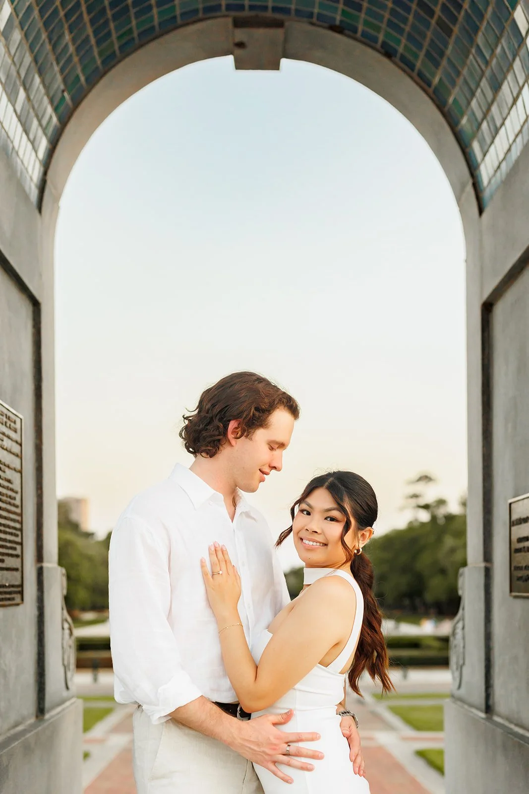 A young couple embracing under an arched structure outdoors, with the woman smiling at the camera and the man looking down at her, during the daytime.
