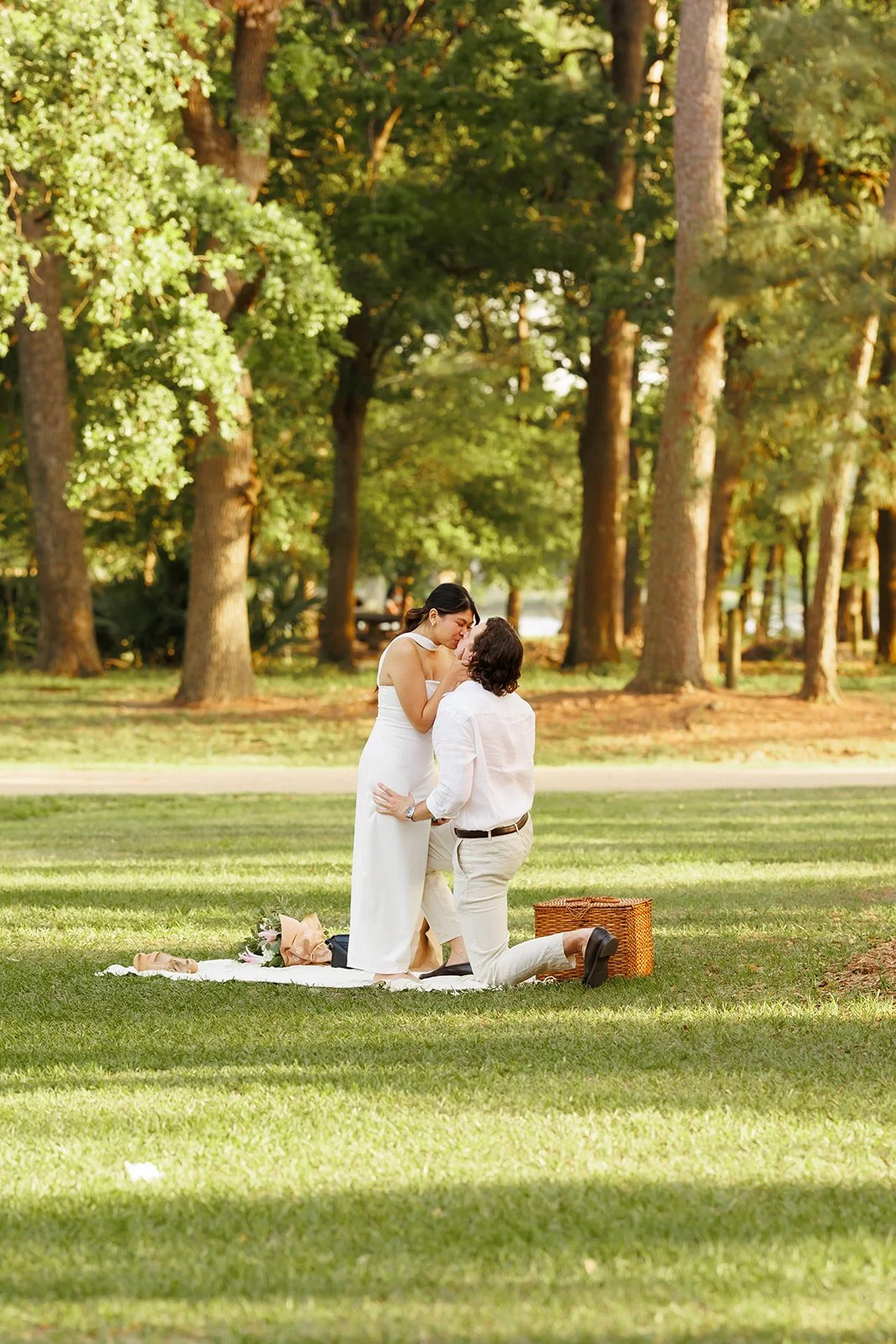 A couple having a wedding moment in a park with tall trees and green grass, with a picnic basket and flowers nearby.