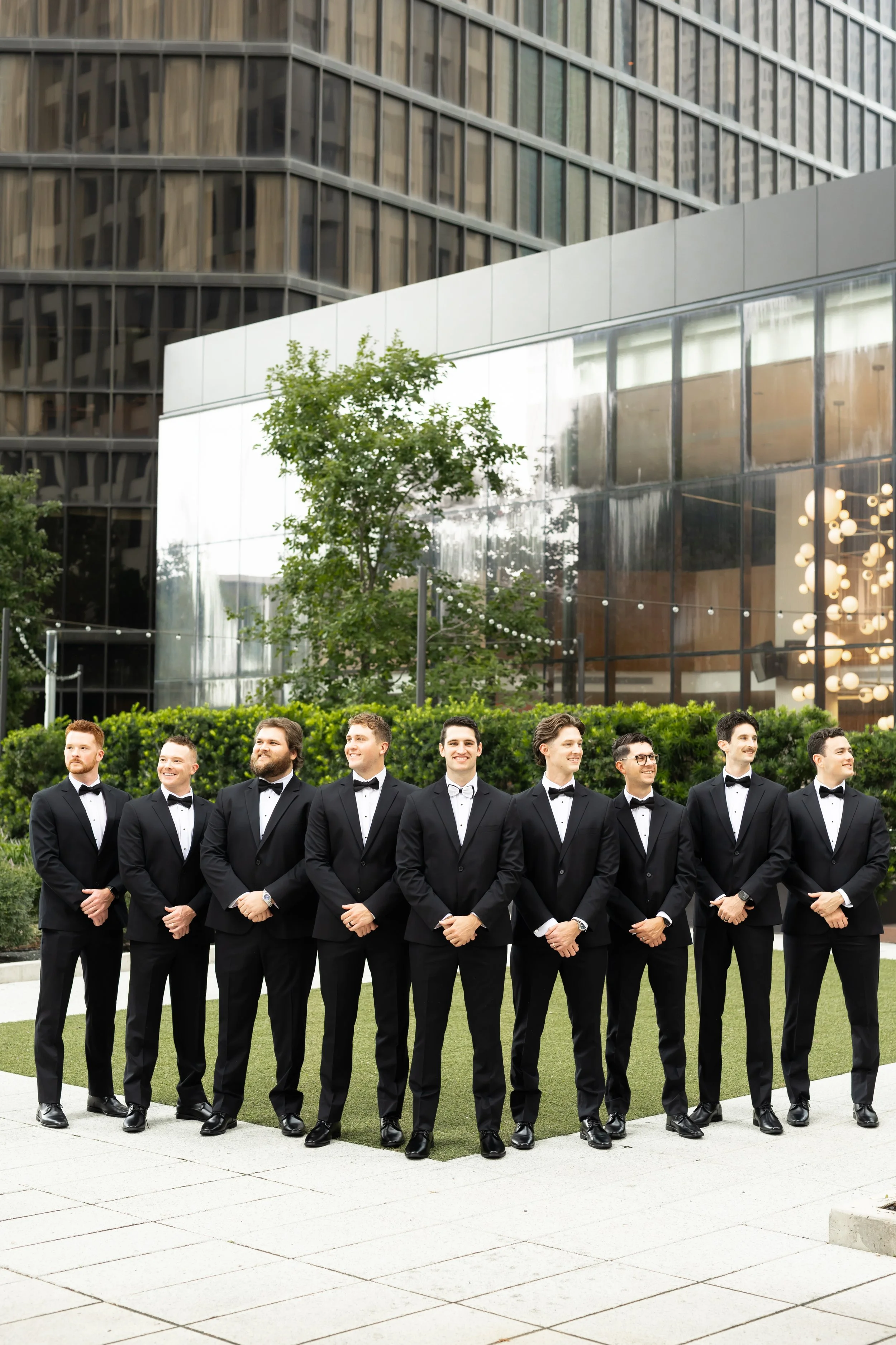 Group of nine men in black tuxedos and bow ties standing outdoors in front of modern glass building during daytime at C Baldwin Hotel by Wild Ivy Creative Photography