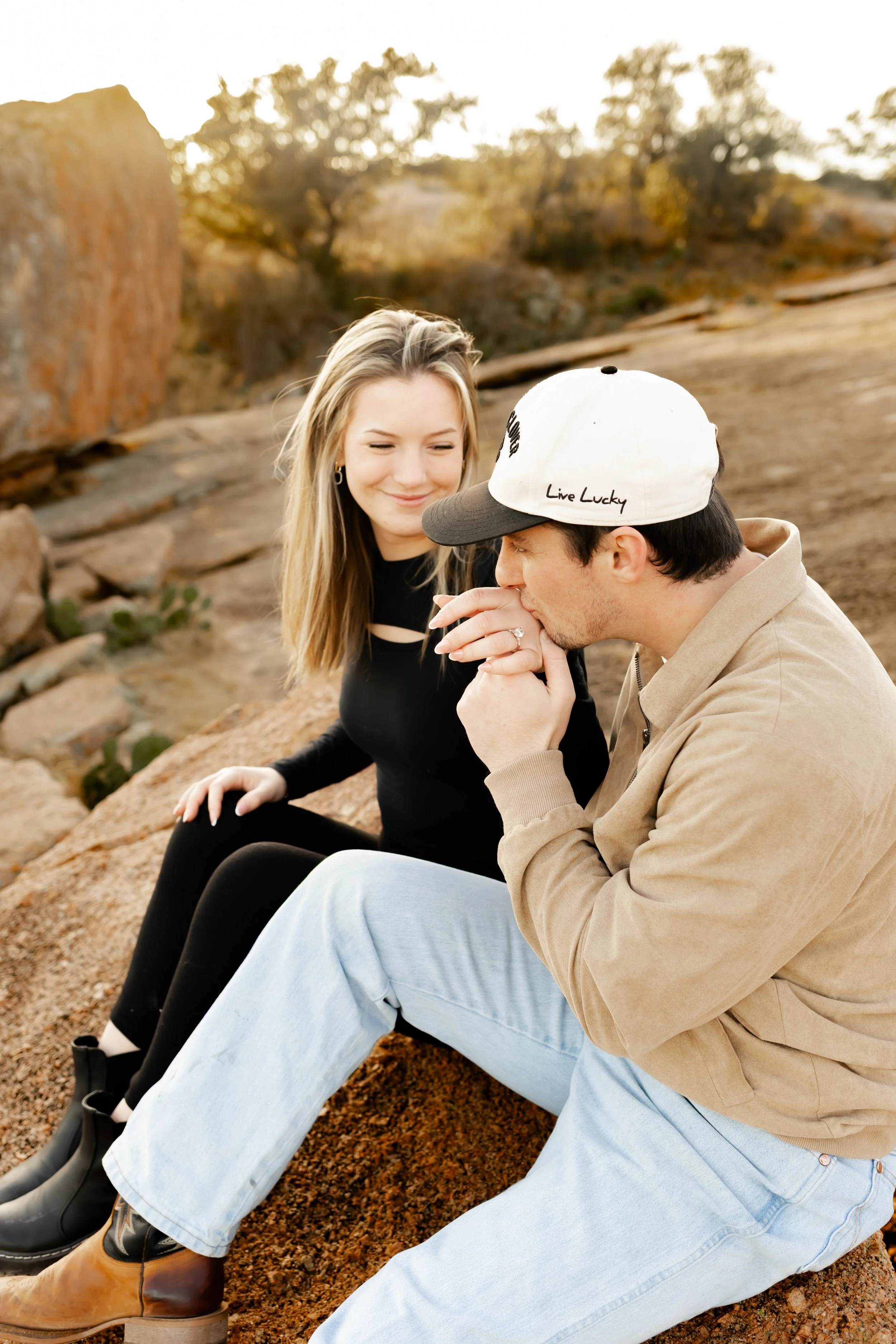 A man kissing a woman on the cheek outdoors with rocks and trees in the background at sunset, by Houston Surprise Proposal Photographer.
