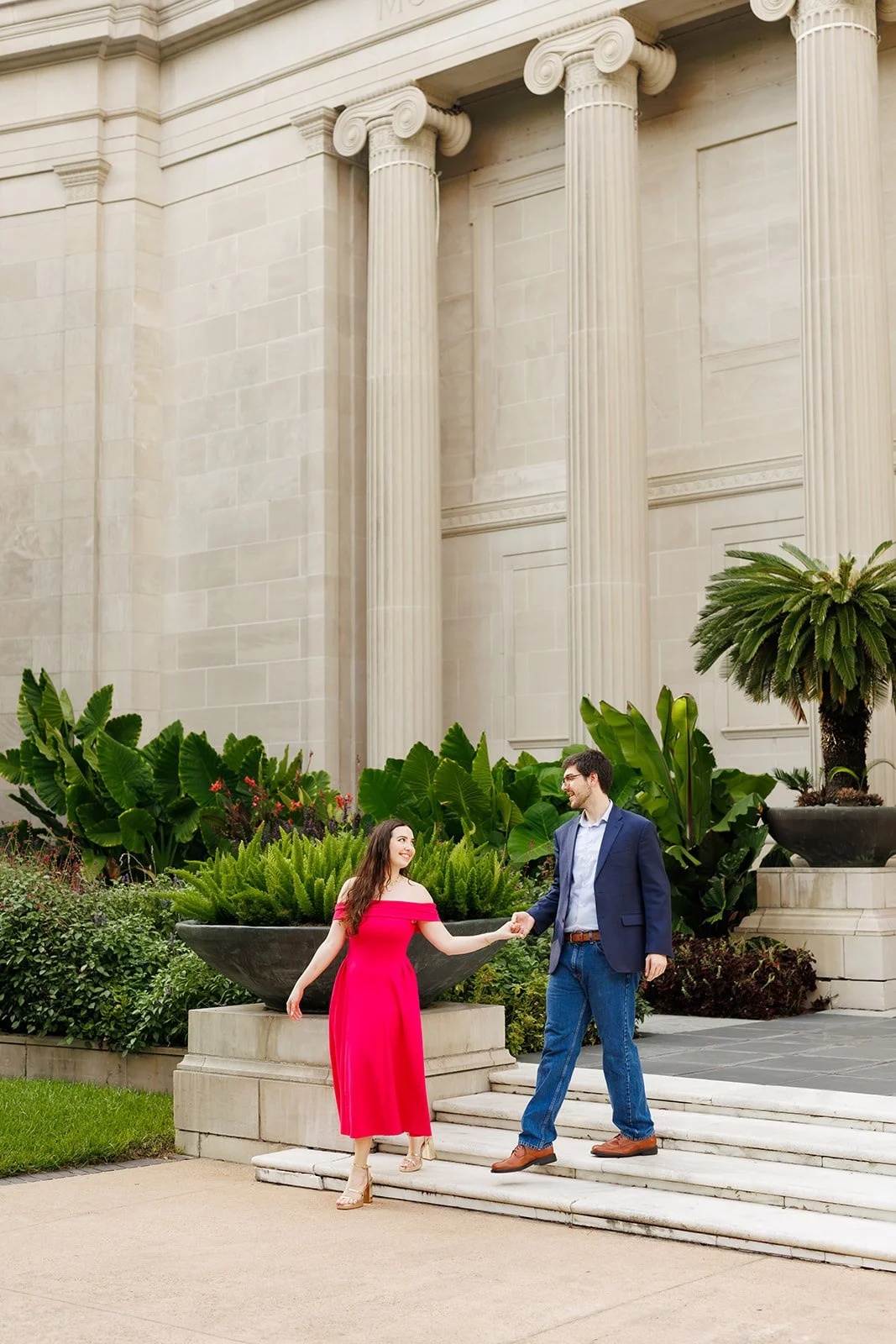 A couple shaking hands on steps outside a building with large columns and lush greenery