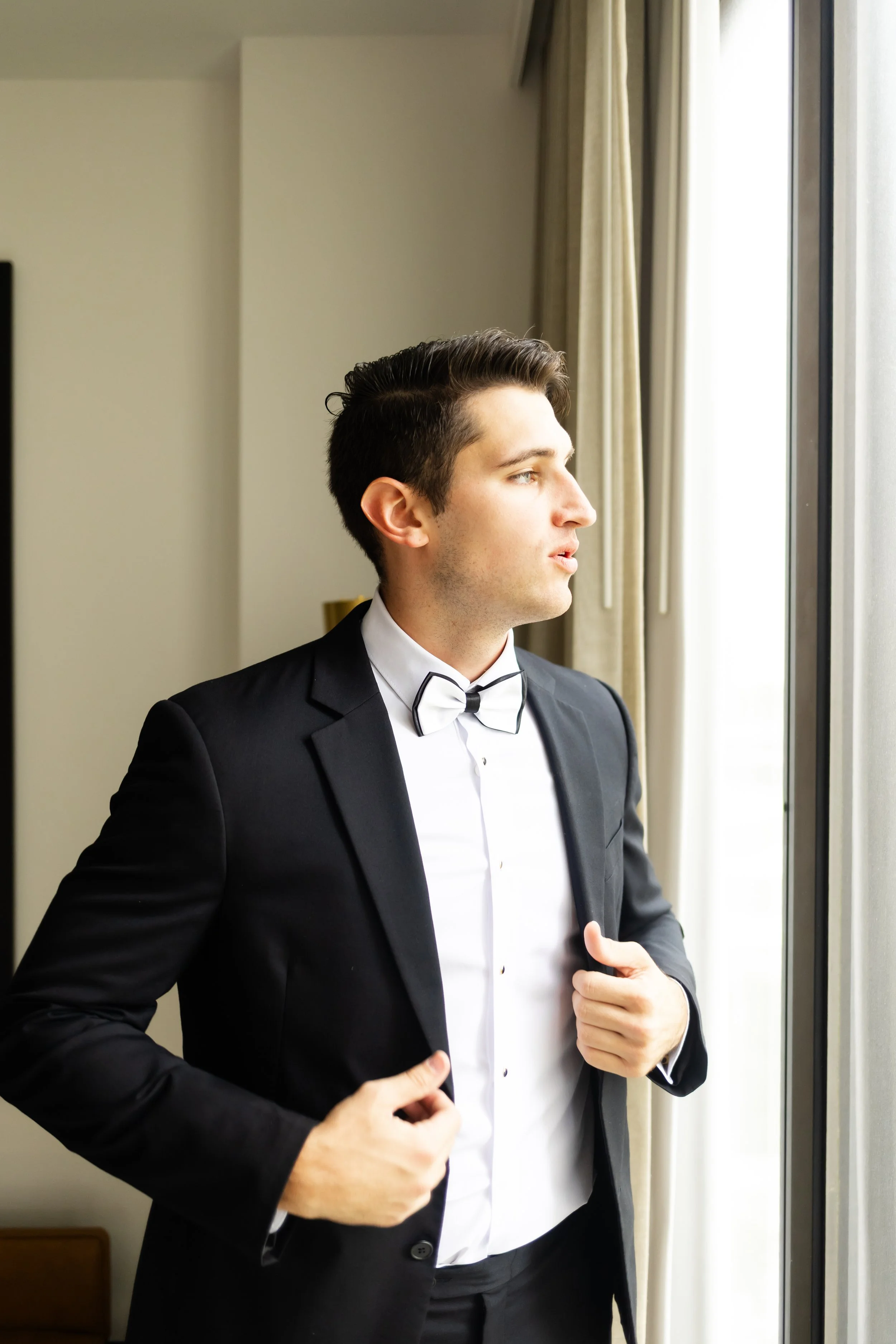 A young man in a tuxedo with a bow tie looking out a window captured by by Wild Ivy Creative Photography