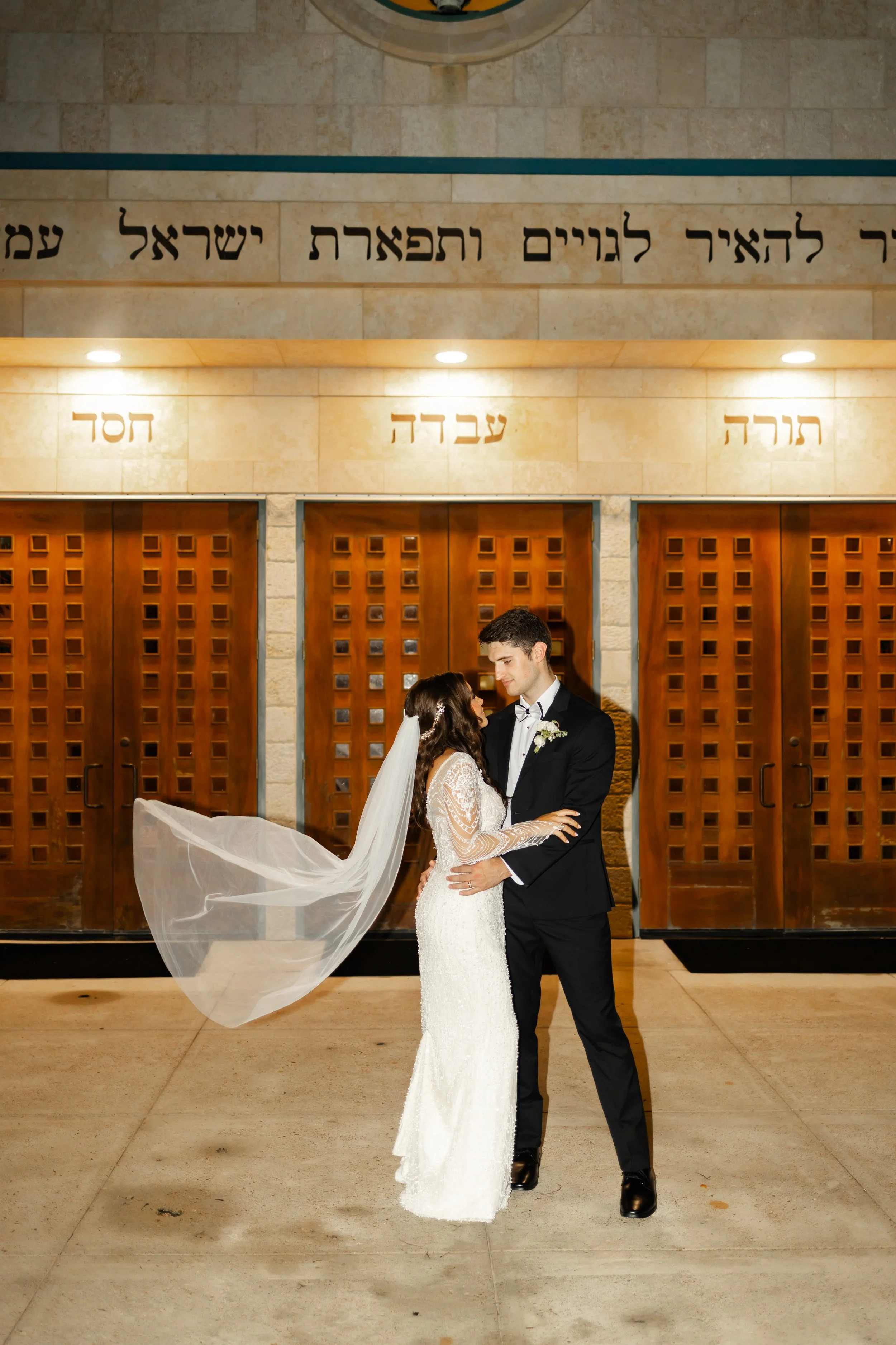 A bride and groom stand close together, gazing into each other's eyes, in front of wooden doors at night. The bride wears a white wedding gown with lace details and a flowing veil, while the groom wears a black suit with a white shirt and boutonniere