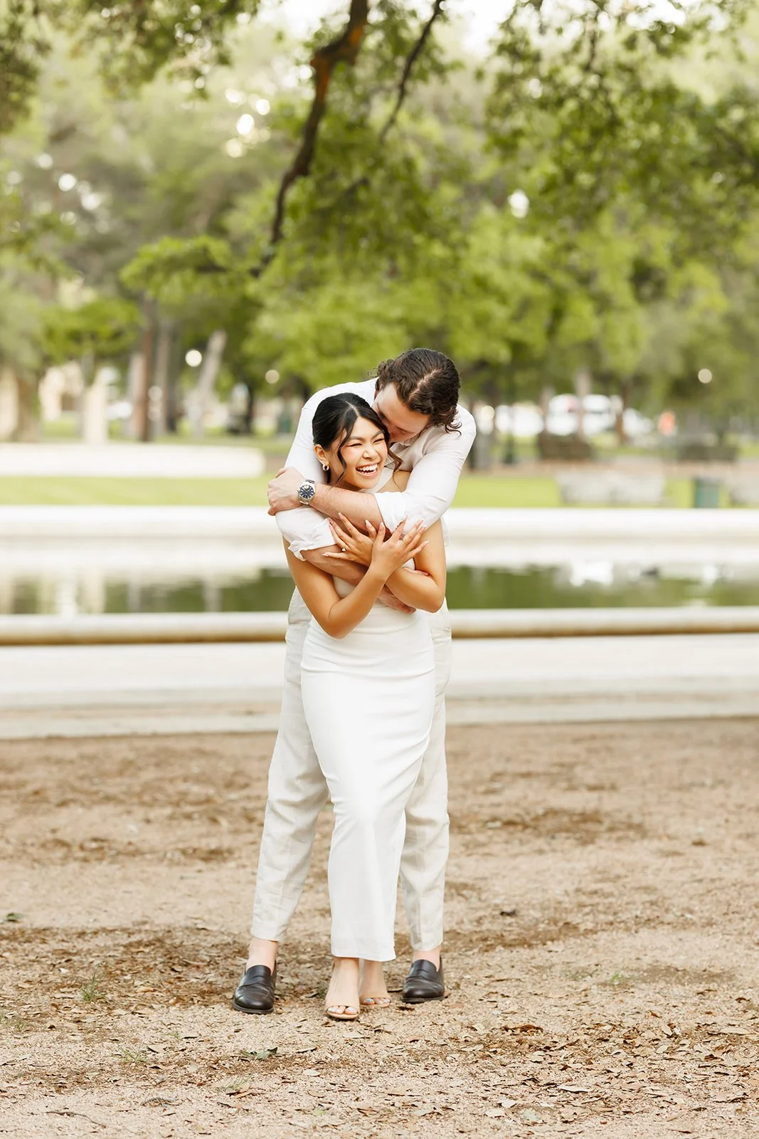 Candid couple’s portrait in Hermann Park in Houston near the water, sharing a joyful embrace and laugh during their engagement shoot.